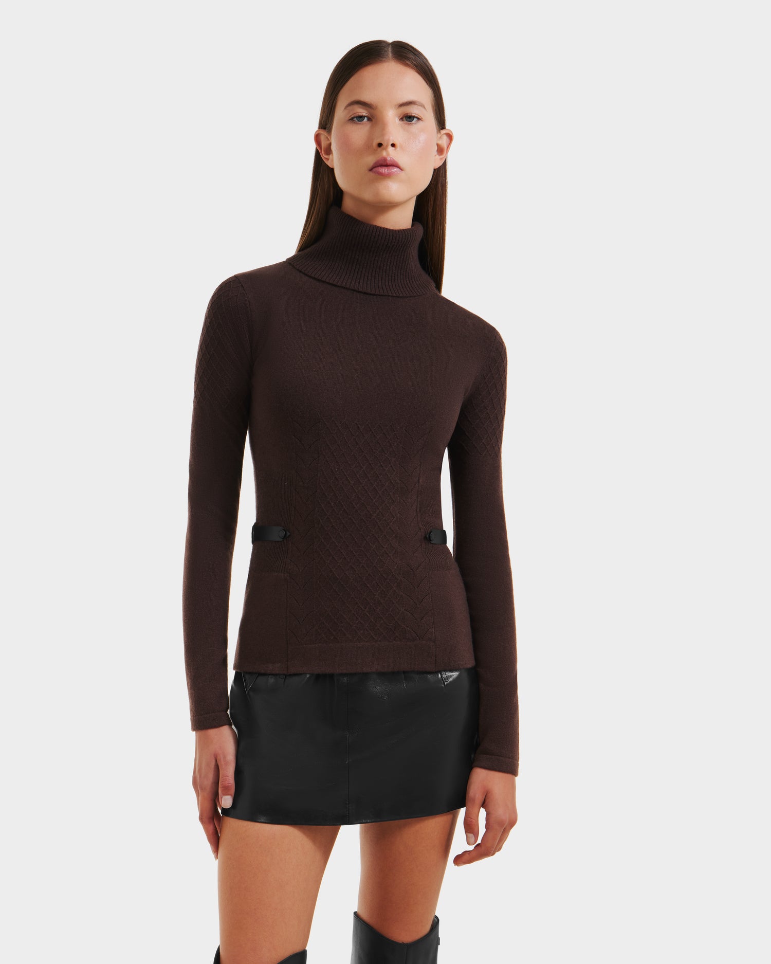 A woman wearing the Rudsak LAURA - 8524596 Mocha knit turtleneck sweater, styled with a black mini skirt and knee-high boots, stands against a plain white background.