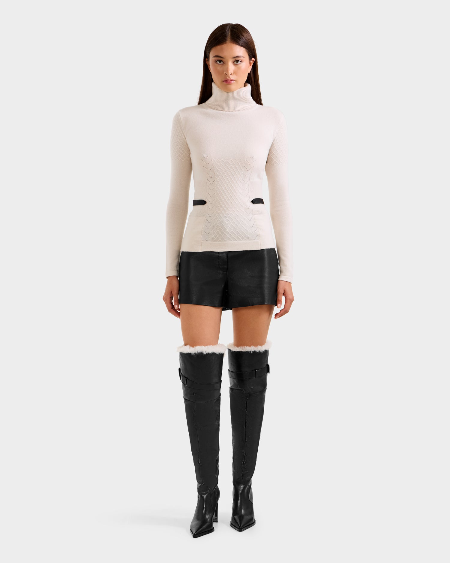 A woman faces forward against a plain background, wearing the Rudsak LAURA - 8524596 Vanilla women's knit turtleneck sweater, styled with black leather shorts and black over-the-knee heeled boots.