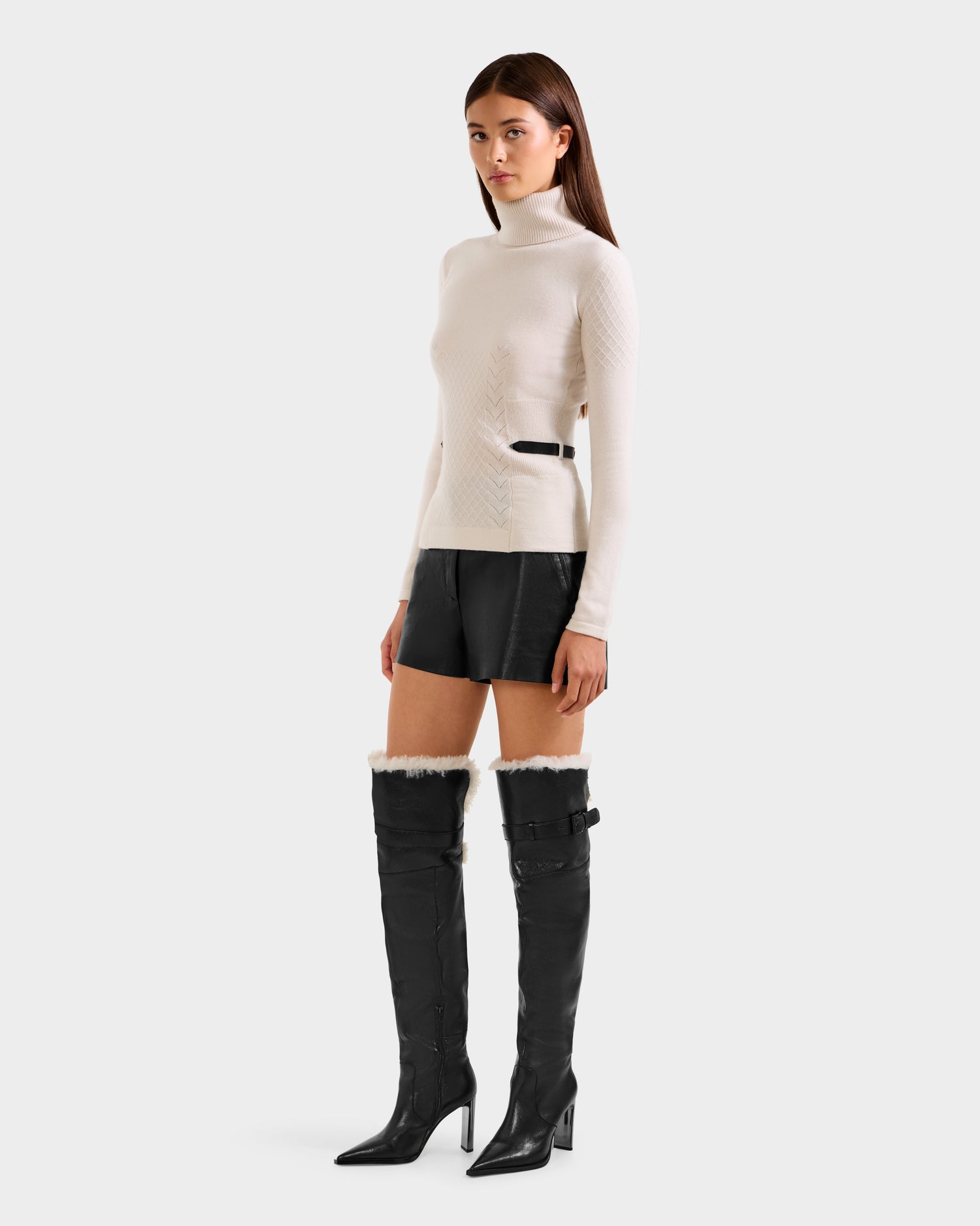 A woman models the Rudsak LAURA - 8524596 Vanilla knit turtleneck sweater, paired with black leather shorts and tall black heeled boots with fur trim and buckles, set against a plain light grey background.