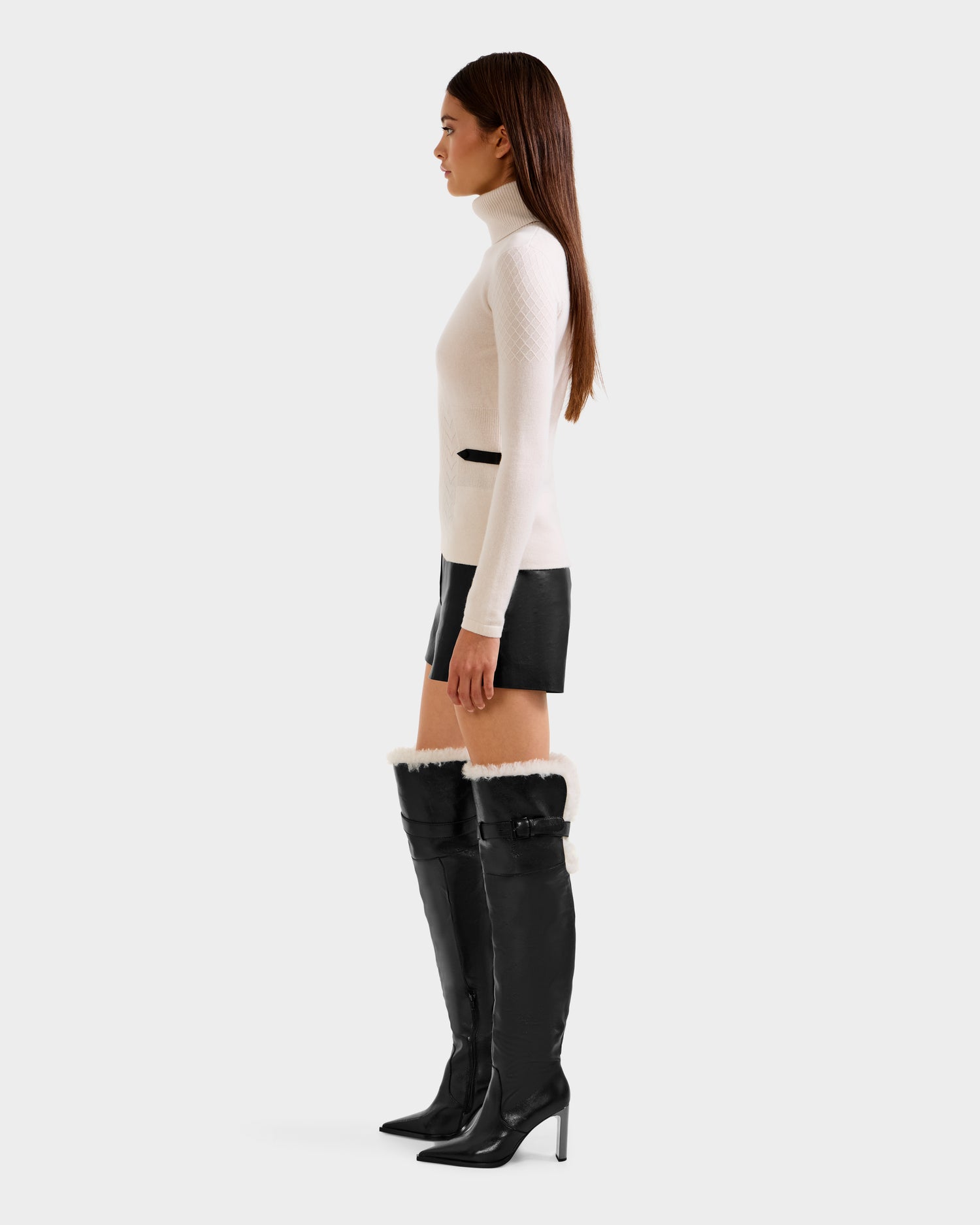 A woman stands in profile against a light gray background, wearing the Rudsak LAURA - 8524596 Vanilla knit turtleneck sweater with black leather shorts and tall black heeled boots with white fur trim.