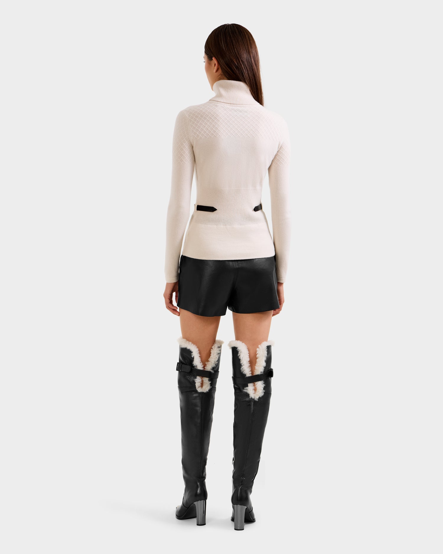 A woman stands facing away, wearing the Rudsak LAURA - 8524596 Vanilla turtleneck sweater with black shorts, tall black boots featuring white fur lining and buckles. Her straight brown hair cascades down her back.
