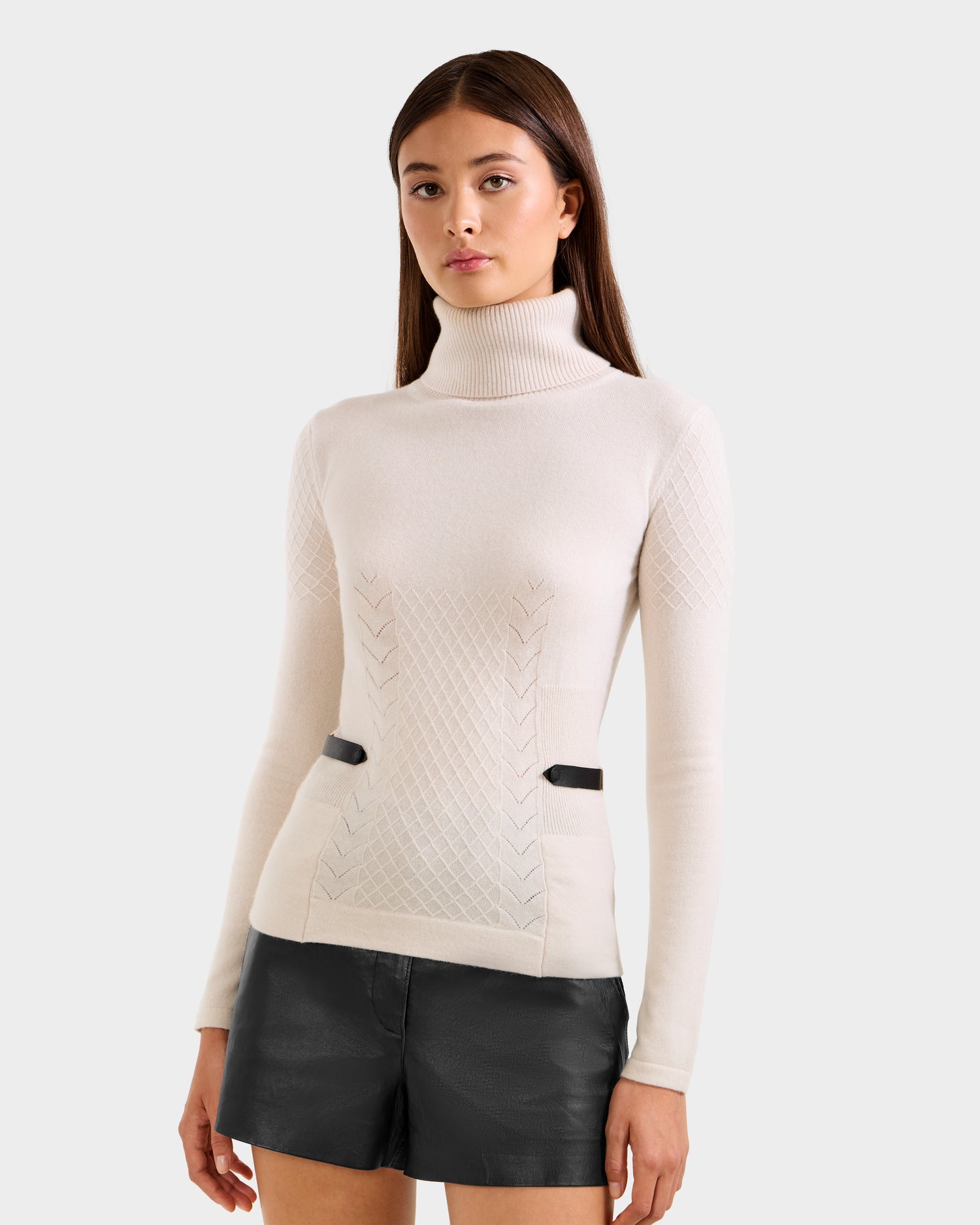 A woman wearing the Rudsak LAURA - 8524596 Vanilla knit turtleneck sweater with textured patterns and black leather shorts stands against a plain, light gray background.