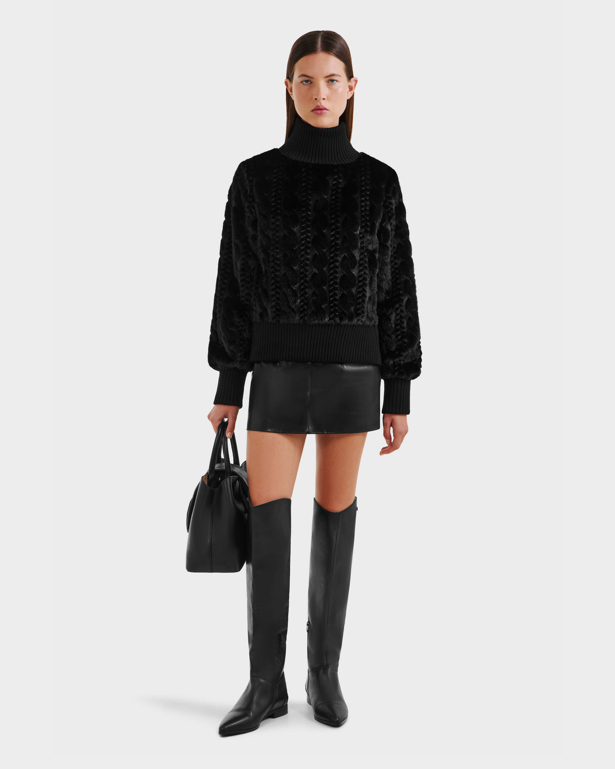 A woman wearing the Rudsak LAYLA F - 8525630 Black cable knit-inspired faux fur sweater, paired with a black leather mini skirt, knee-high black boots, and holding a black handbag, stands against a plain background.