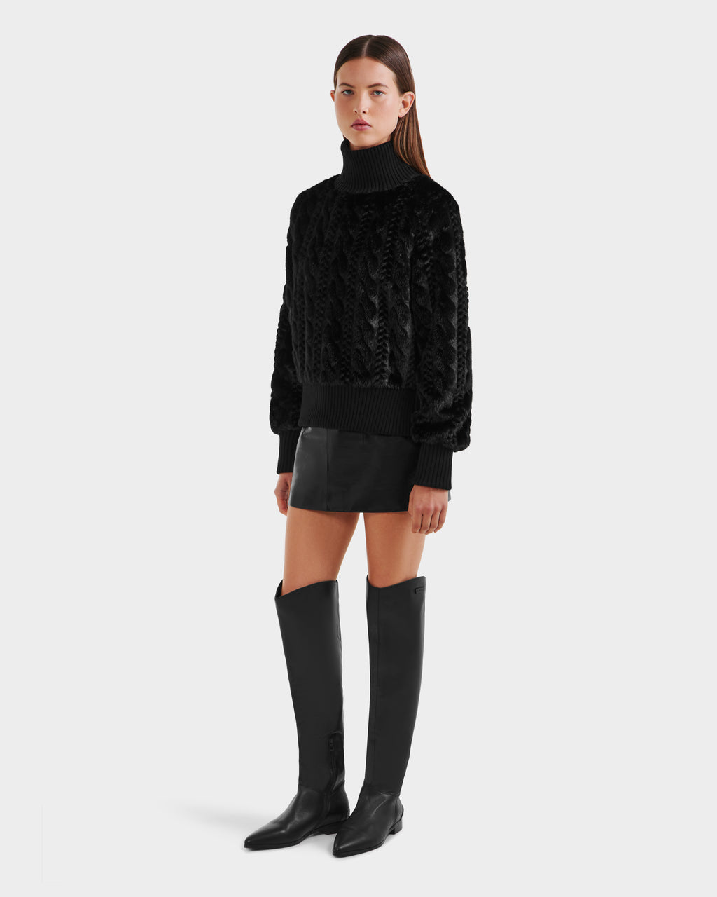A woman with straight, shoulder-length hair wears the Rudsak LAYLA F - 8525630 Black faux fur turtleneck sweater, paired with a black mini skirt and knee-high boots—an ideal look for après-ski style.