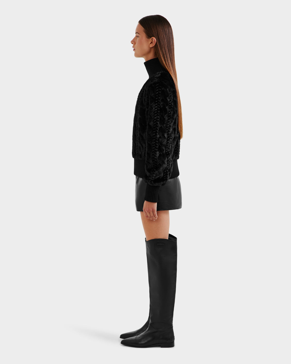 A woman in profile poses against a light background, wearing the LAYLA F - 8525630 Black faux fur turtleneck by Rudsak, a black leather mini skirt, and knee-high black boots. Her straight brown hair falls past her shoulders.
