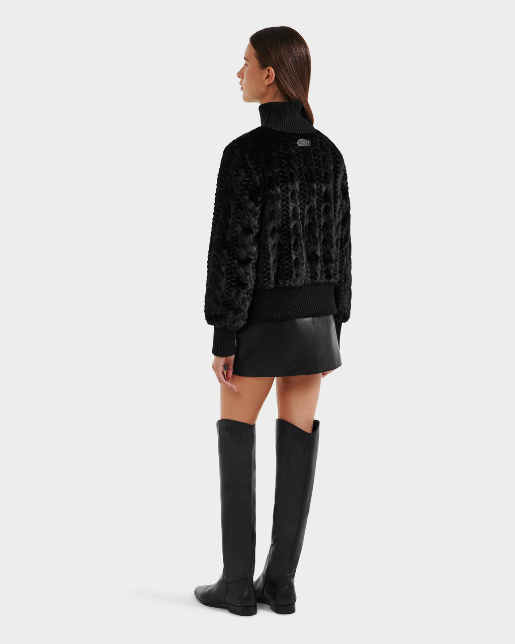 A woman faces away, wearing the Rudsak LAYLA F - 8525630 Black faux fur jacket with a black leather mini skirt and knee-high boots. Her straight brown hair is down, posing against a plain light gray background.
