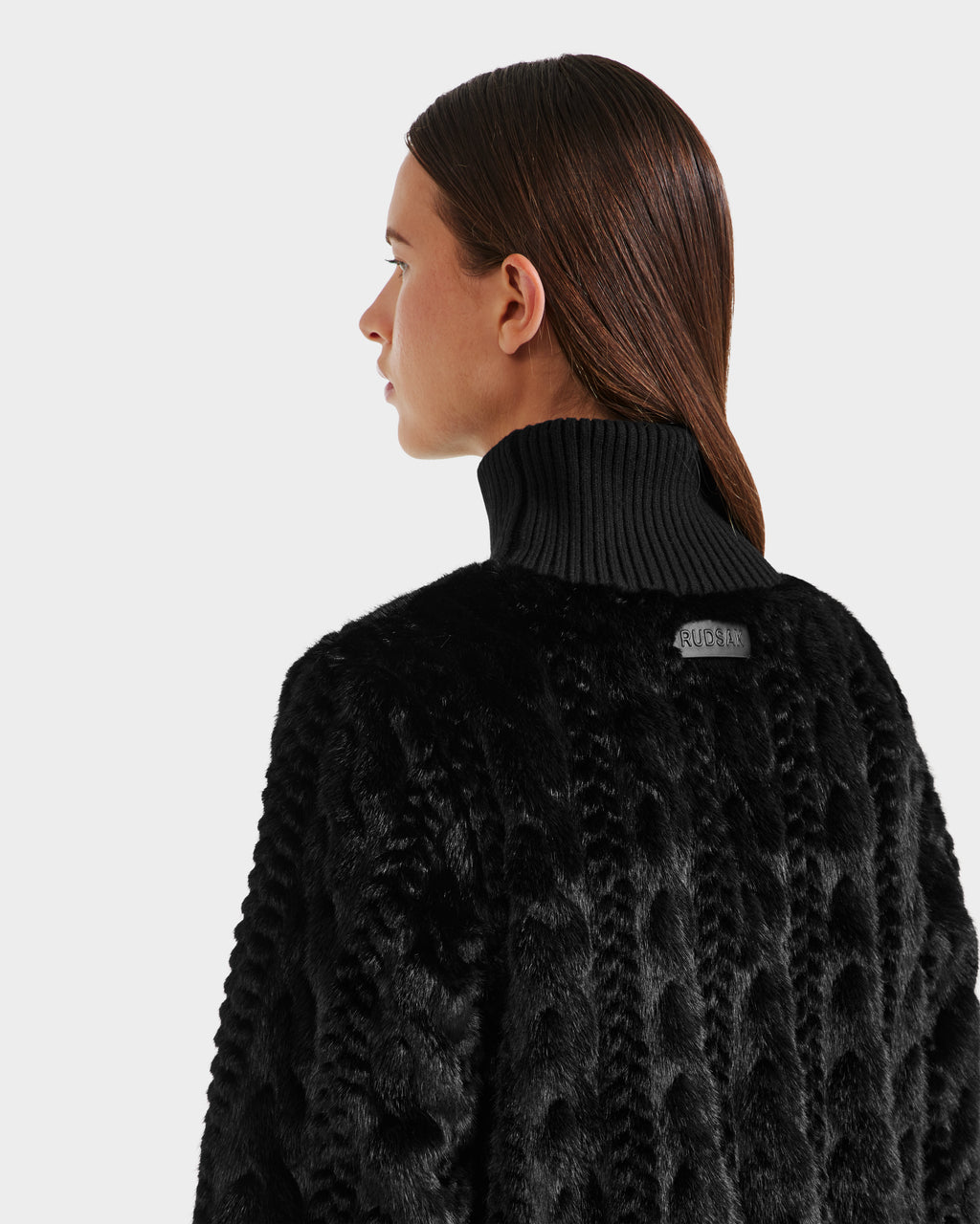 Woman with straight brown hair wears the Rudsak LAYLA F - 8525630 Black cable knit-inspired faux fur coat, featuring a high ribbed collar, shown from the back and looking left against a plain light background.