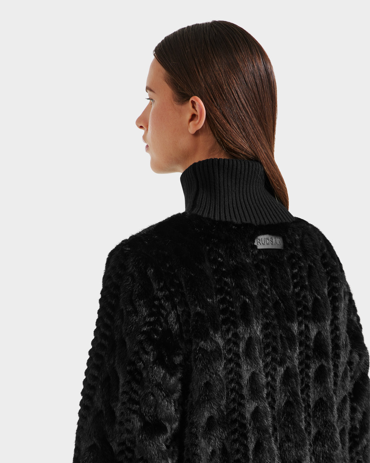 Woman with straight brown hair wears the Rudsak LAYLA F - 8525630 Black cable knit-inspired faux fur coat, featuring a high ribbed collar, shown from the back and looking left against a plain light background.