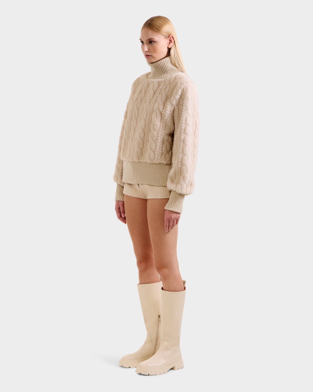 A woman in Rudsak’s LAYLA F - 8525630 Vanilla sweater stands before a plain backdrop wearing beige shorts and cream knee-high boots, her neutral expression capturing effortless après-ski style.