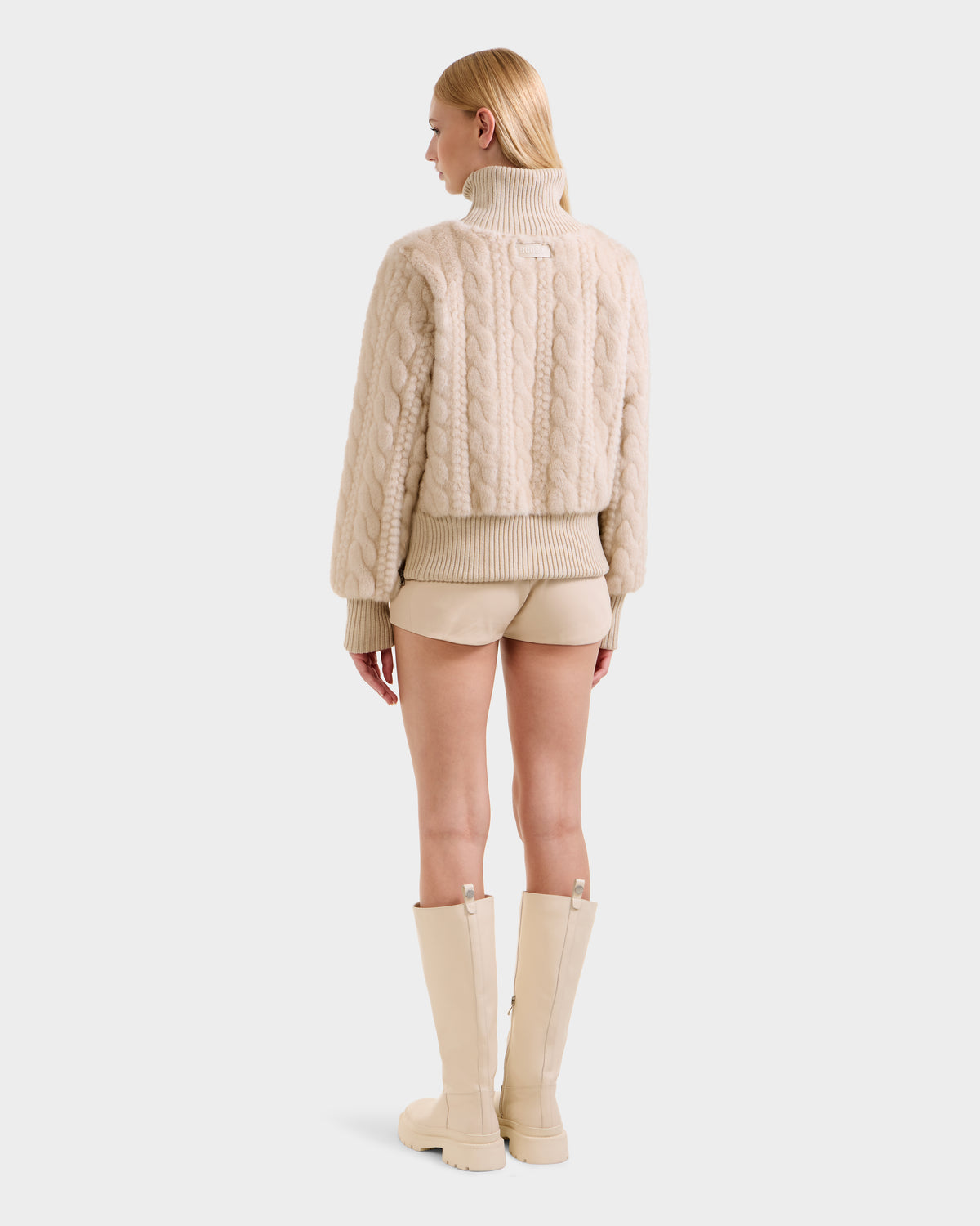 A woman with straight blonde hair stands facing away against a white background, wearing the Rudsak LAYLA F - 8525630 Vanilla cable-knit turtleneck sweater, beige shorts, and knee-high beige boots.