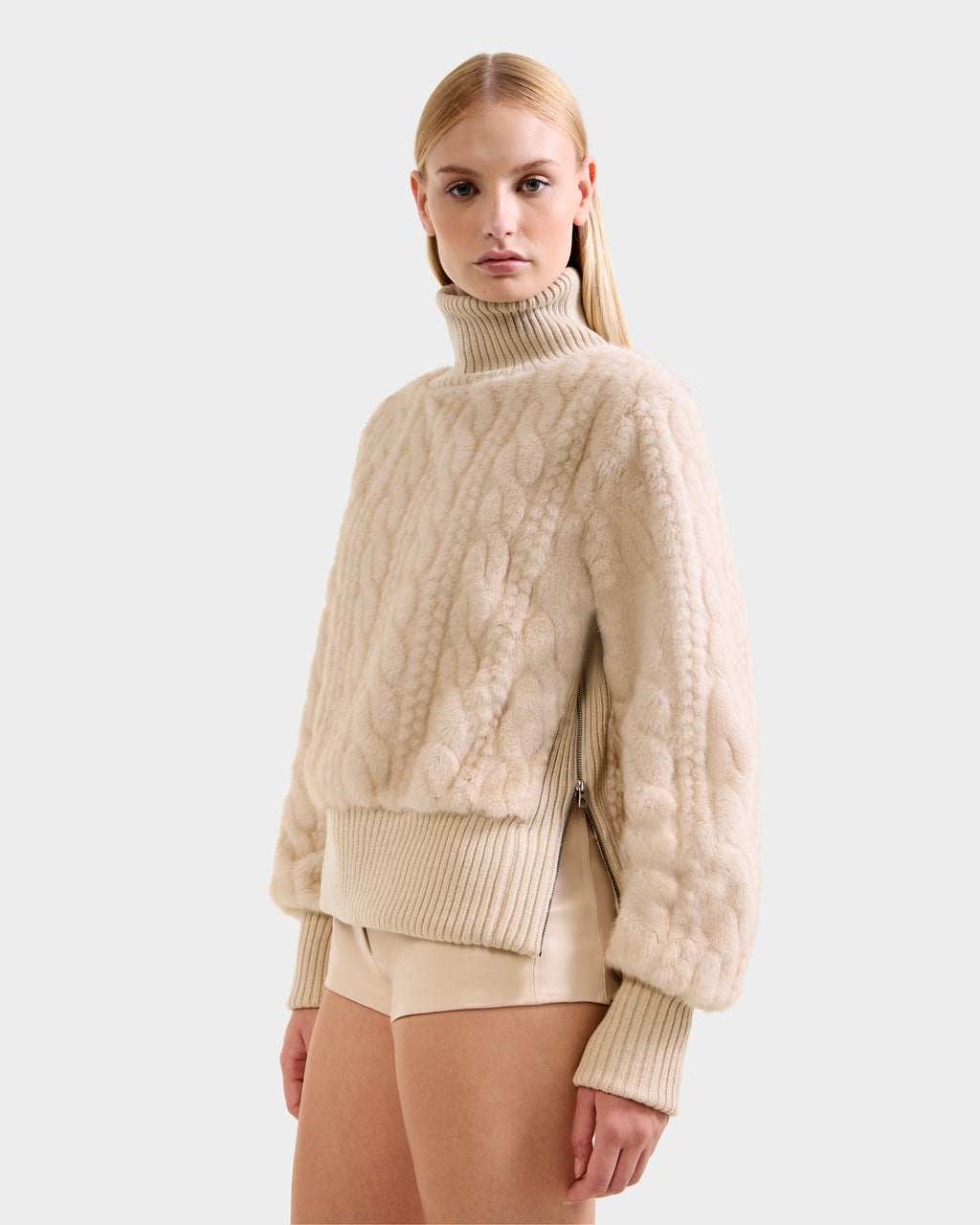A woman with straight blonde hair wears the Rudsak LAYLA F - 8525630 Vanilla chunky cable knit-inspired turtleneck sweater with ribbed cuffs and hem, paired with beige shorts, standing against a light gray background.