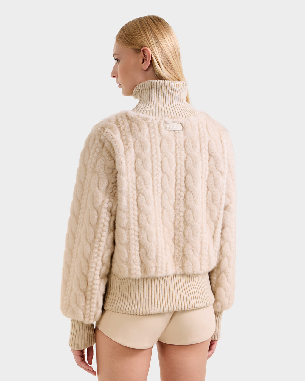 A woman with straight blonde hair models the Rudsak LAYLA F - 8525630 Vanilla, a chunky, cream cable knit-inspired faux fur turtleneck sweater, paired with beige shorts. She is turned away, displaying the back against a plain light background.