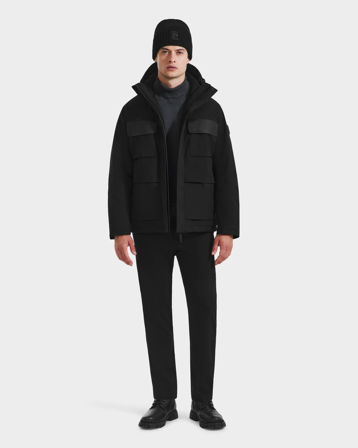 A man faces forward against a white background, wearing the Rudsak LAZIO - 6125527 Black men’s parka with down and feather fill, paired with a black beanie, dark sweater, black pants, and shoes.
