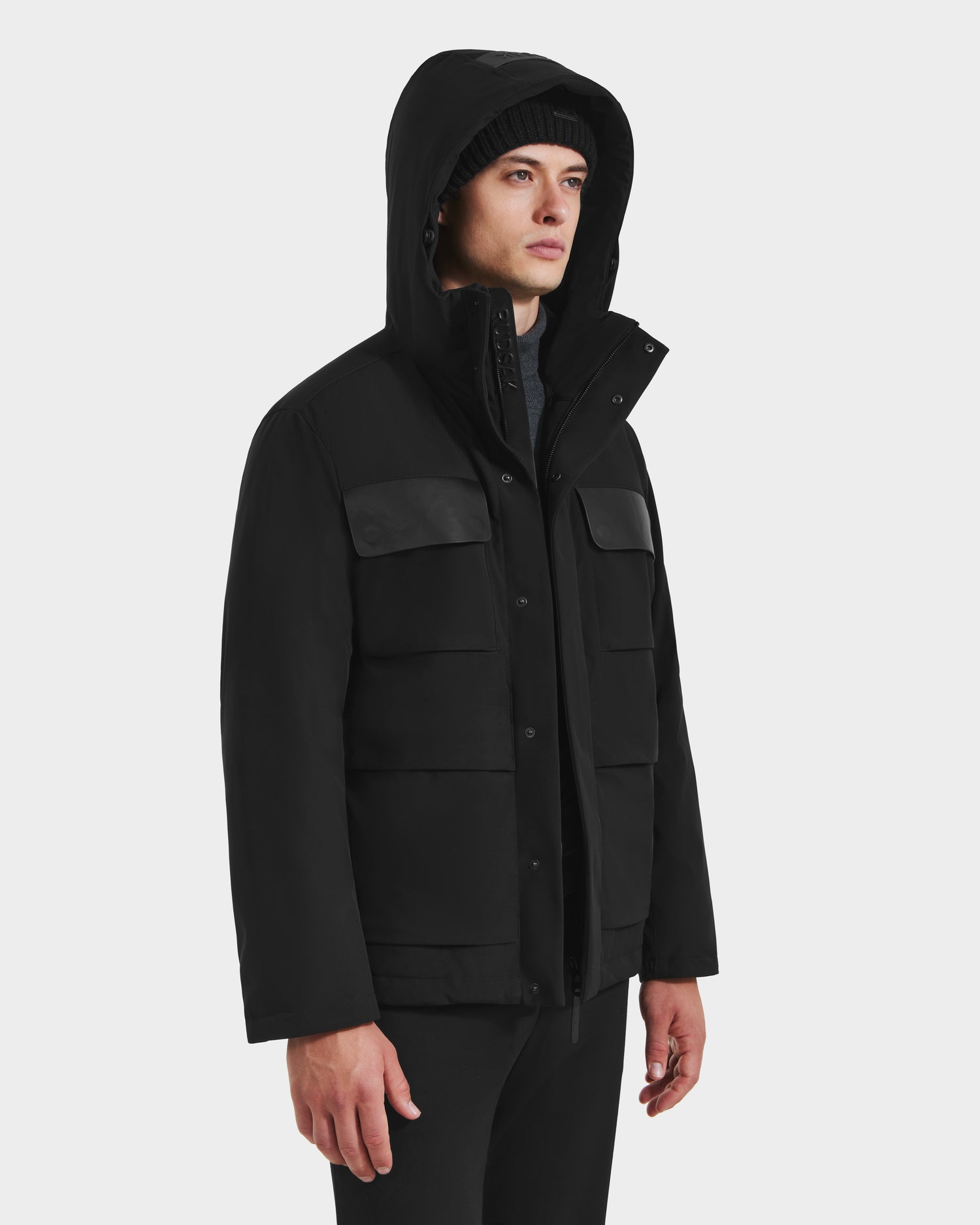 A person in the Rudsak LAZIO - 6125527 Black men’s parka, featuring front pockets and down-feather fill, stands against a plain white background in a slightly turned pose—a true winter essential.