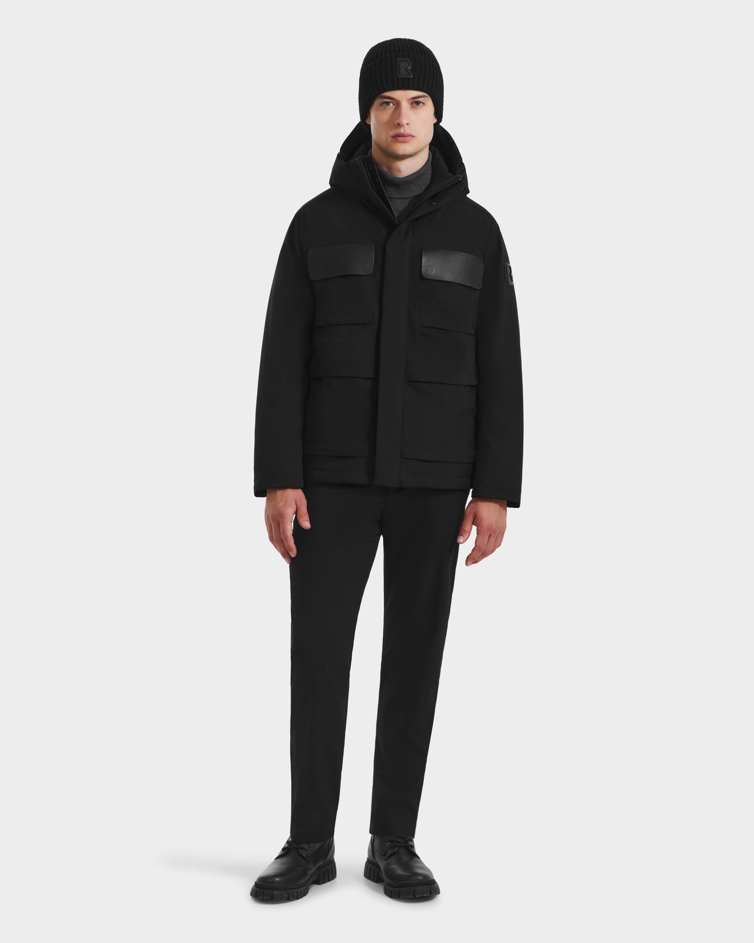 A man faces forward wearing the Rudsak LAZIO - 6125527 Black mens parka with four front pockets, paired with a black beanie, pants, and shoes—an understated winter essential—set against a plain white background.