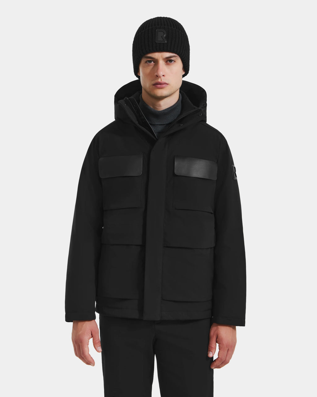 A person wearing the Rudsak LAZIO - 6125527 Black down insulated jacket with four front pockets and a hood, black knit beanie, and black pants stands facing forward against a plain light background.