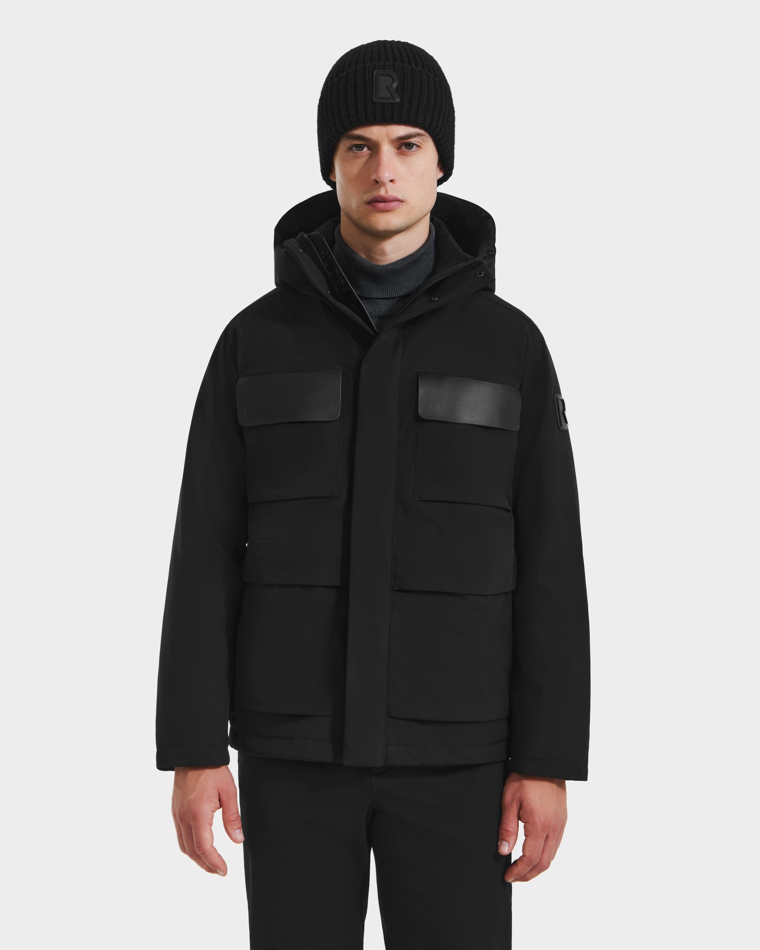 A person wearing the Rudsak LAZIO - 6125527 Black down insulated jacket with four front pockets and a hood, black knit beanie, and black pants stands facing forward against a plain light background.