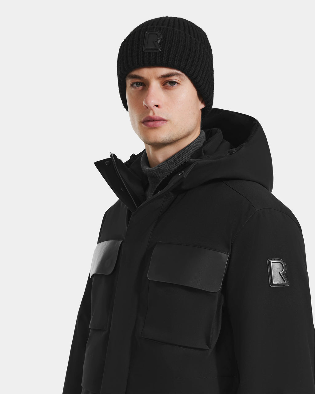 A person wears the Rudsak LAZIO - 6125527 Black men’s parka with large front pockets and a black knit beanie, both featuring a bold R logo, standing against a plain light background—perfect winter essentials.