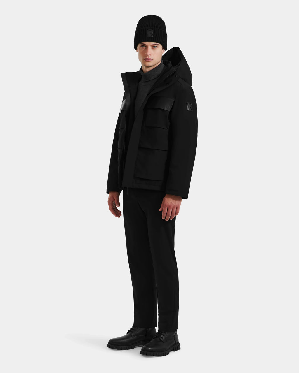 A person stands slightly turned left, wearing the Rudsak LAZIO - 6125527 Black down jacket, paired with a black beanie, pants, and shoes, set against a plain light background.