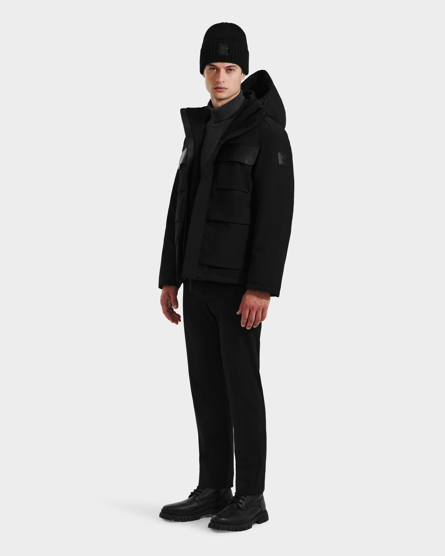 A person stands slightly turned left, wearing the Rudsak LAZIO - 6125527 Black down jacket, paired with a black beanie, pants, and shoes, set against a plain light background.