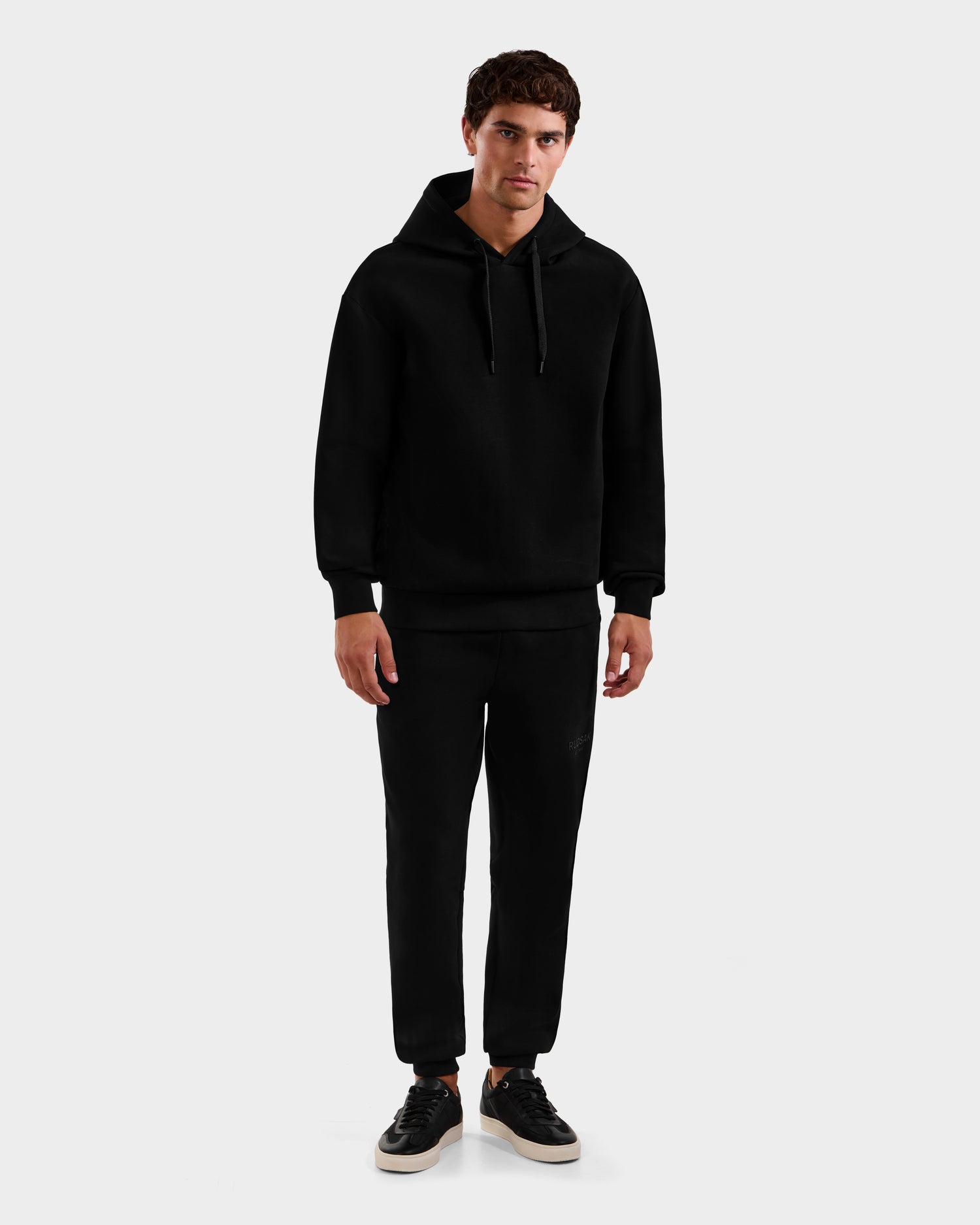 A man faces forward against a light gray background, wearing Rudsak’s LEROY - U525509 Black M unisex hoodie and jogger pants in luxe cotton-blend, paired with black sneakers with white soles.