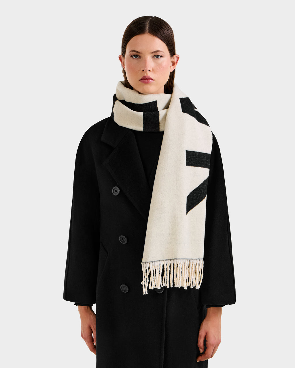 A woman wears the Rudsak LEV - U425506 Black long coat and a thick cream reversible unisex scarf with black patterns and fringed ends, posing against a plain light gray background—an elegant cold-weather look.