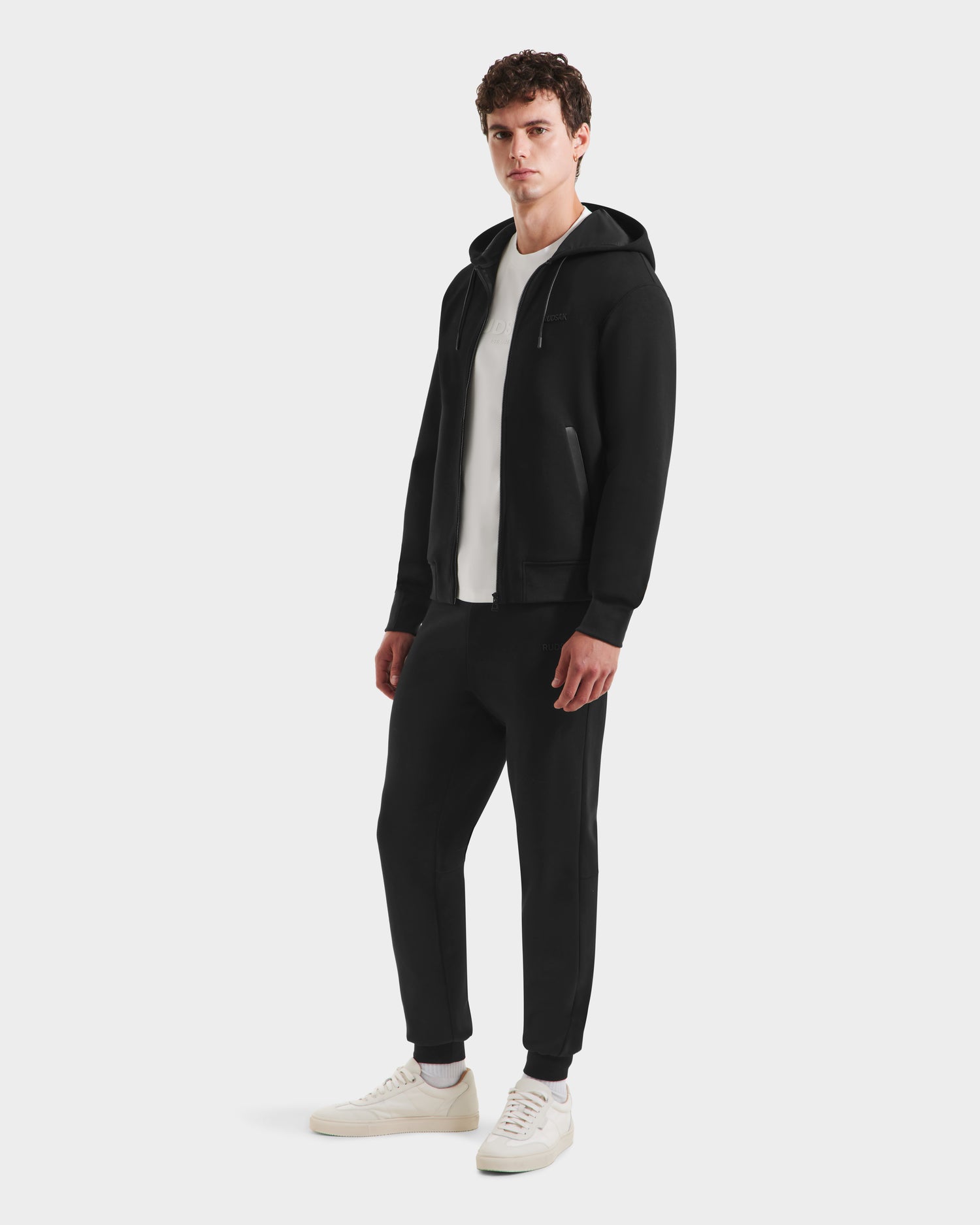 A man wears the Rudsak LEVI - 9525552 Black M unisex zip-up hoodie with vegan leather trims, matching joggers, a white tee, and sneakers against a plain background—an effortless elevated basics look.