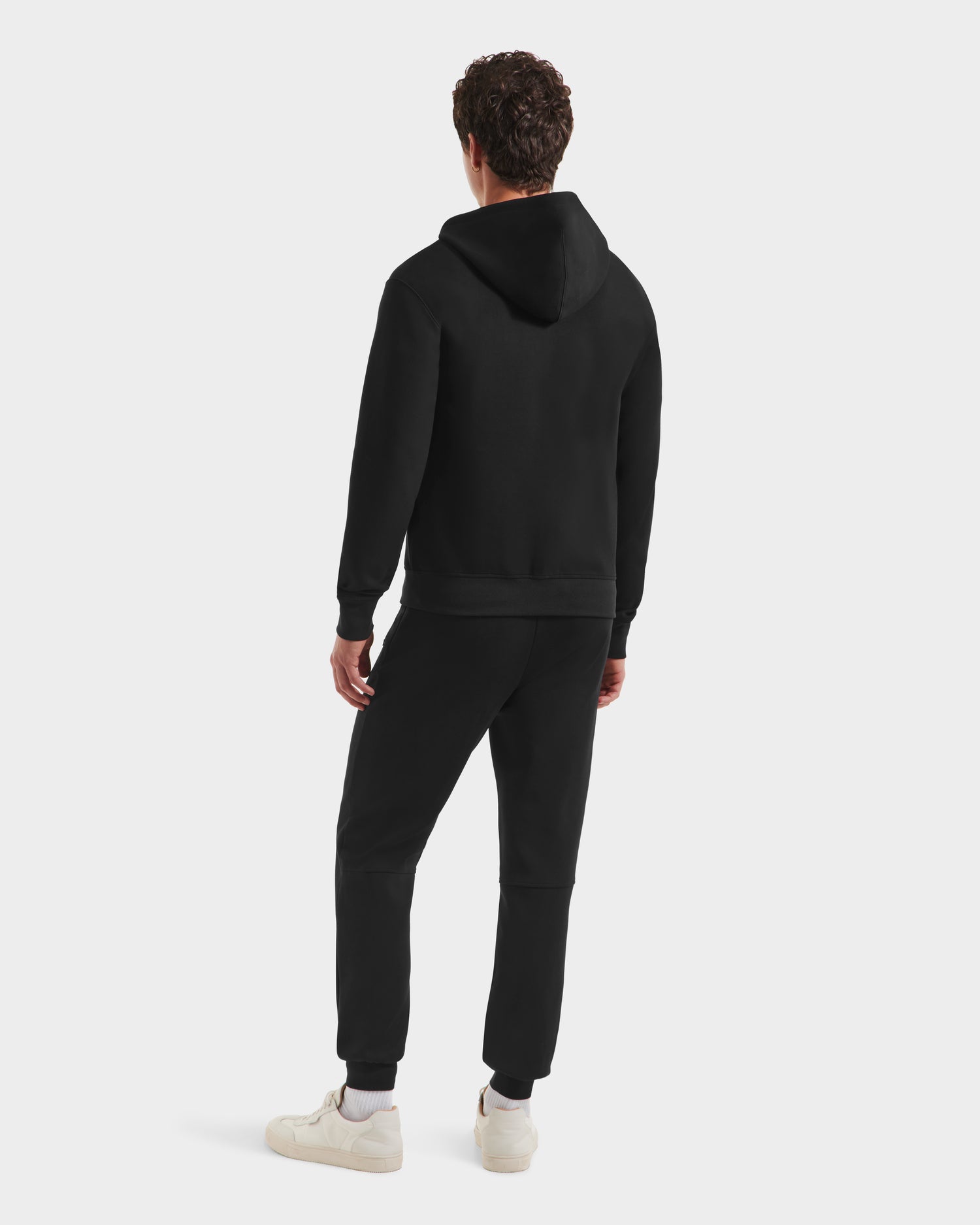 A man, seen from behind, wears the Rudsak LEVI - 9525552 Black M unisex zip-up hoodie, paired with black jogger pants and white sneakers, exemplifying modern elevated basics against a plain white background.