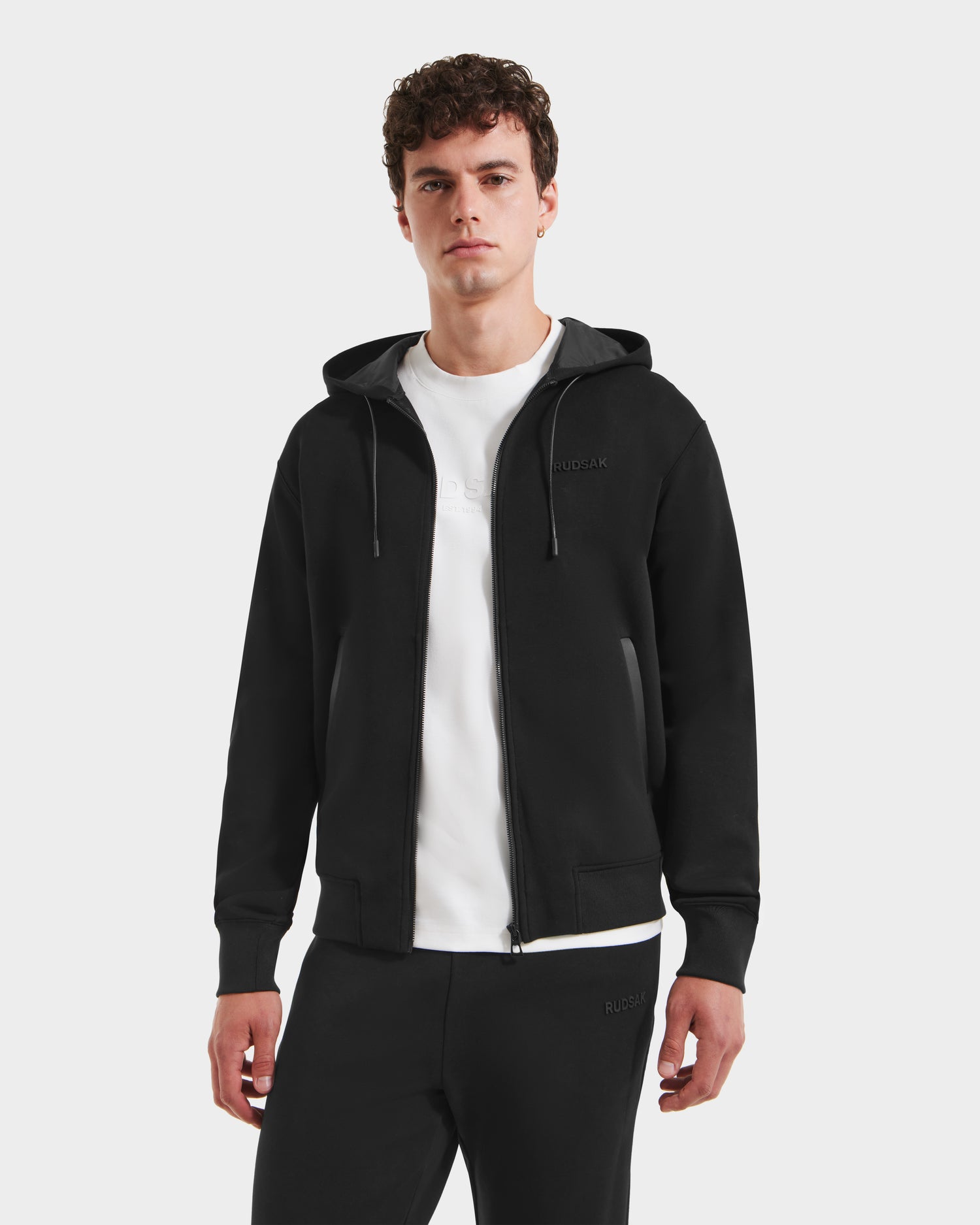 A young man with short curly hair wears the Rudsak LEVI - 9525552 Black M unisex zip-up hoodie over a white T-shirt and black pants, standing facing forward against a plain white background.