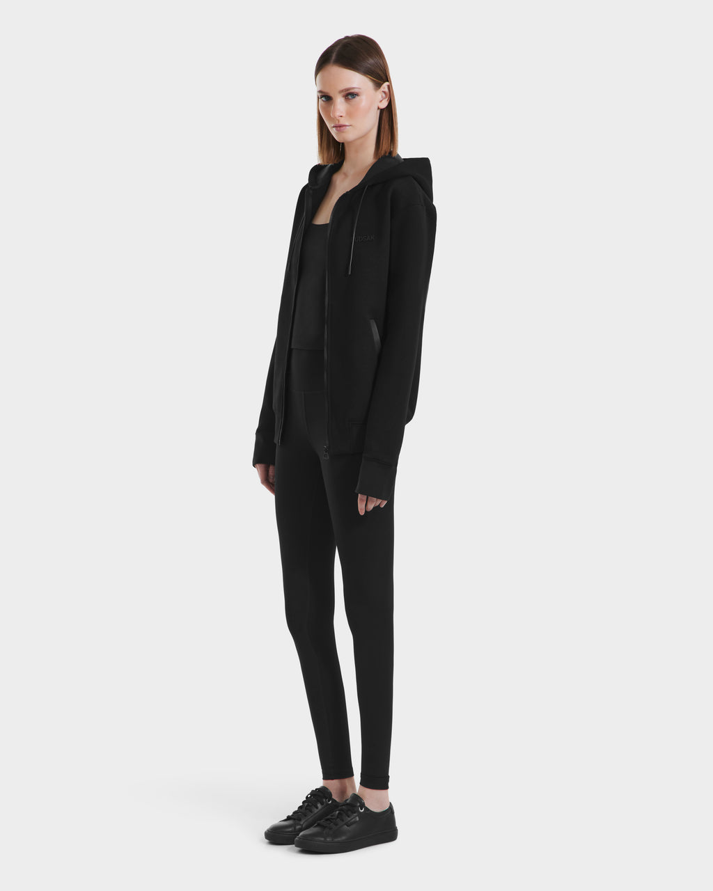 A woman with straight brown hair and a neutral expression stands against a white background, wearing Rudsaks LEVI - 9525552 Black W unisex zip-up hoodie with black fitted pants and sneakers, her hands by her sides.