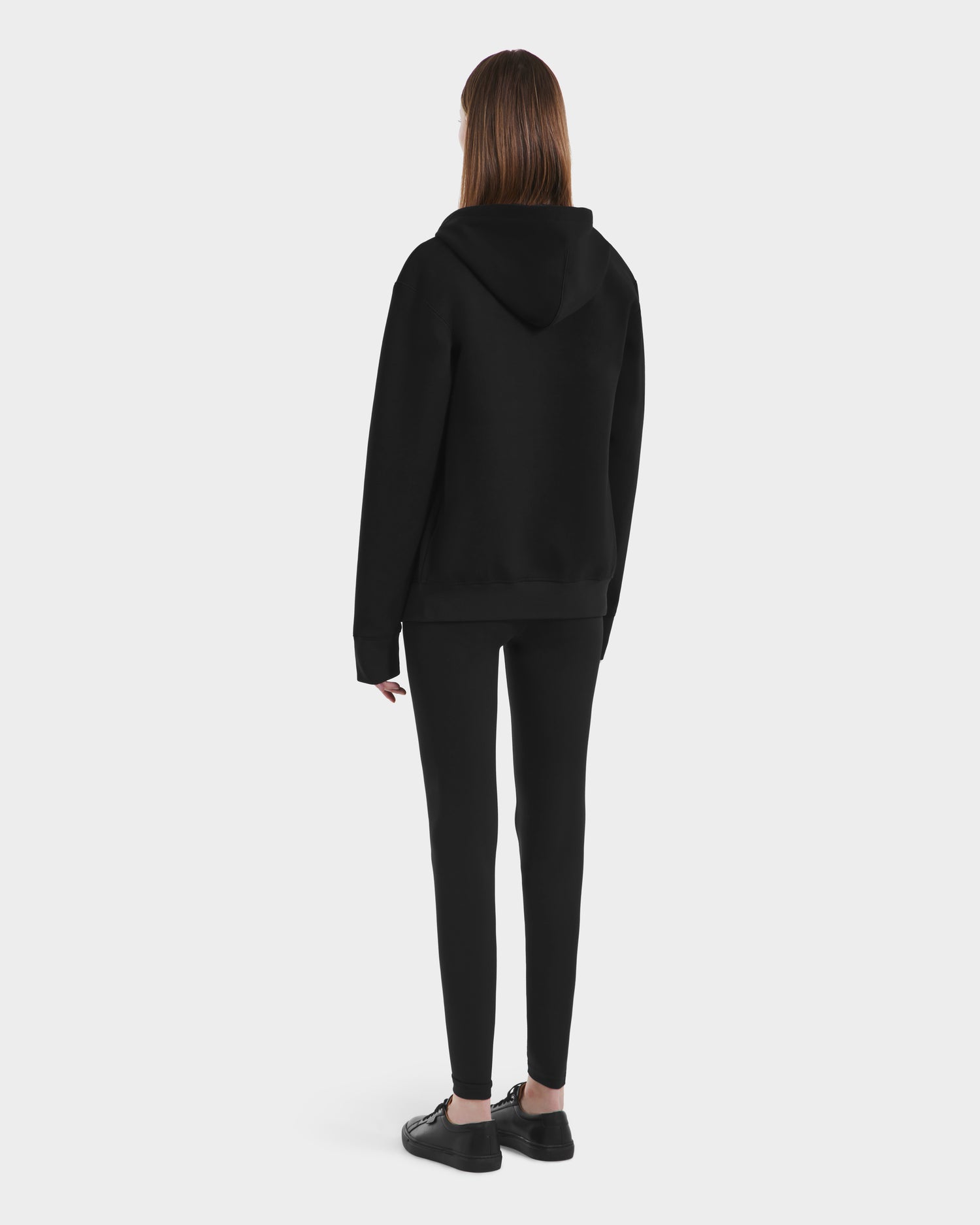 A person with straight brown hair wears the Rudsak LEVI - 9525552 Black W unisex zip-up hoodie, black leggings, and black shoes, standing with their back to the camera against a plain white background.