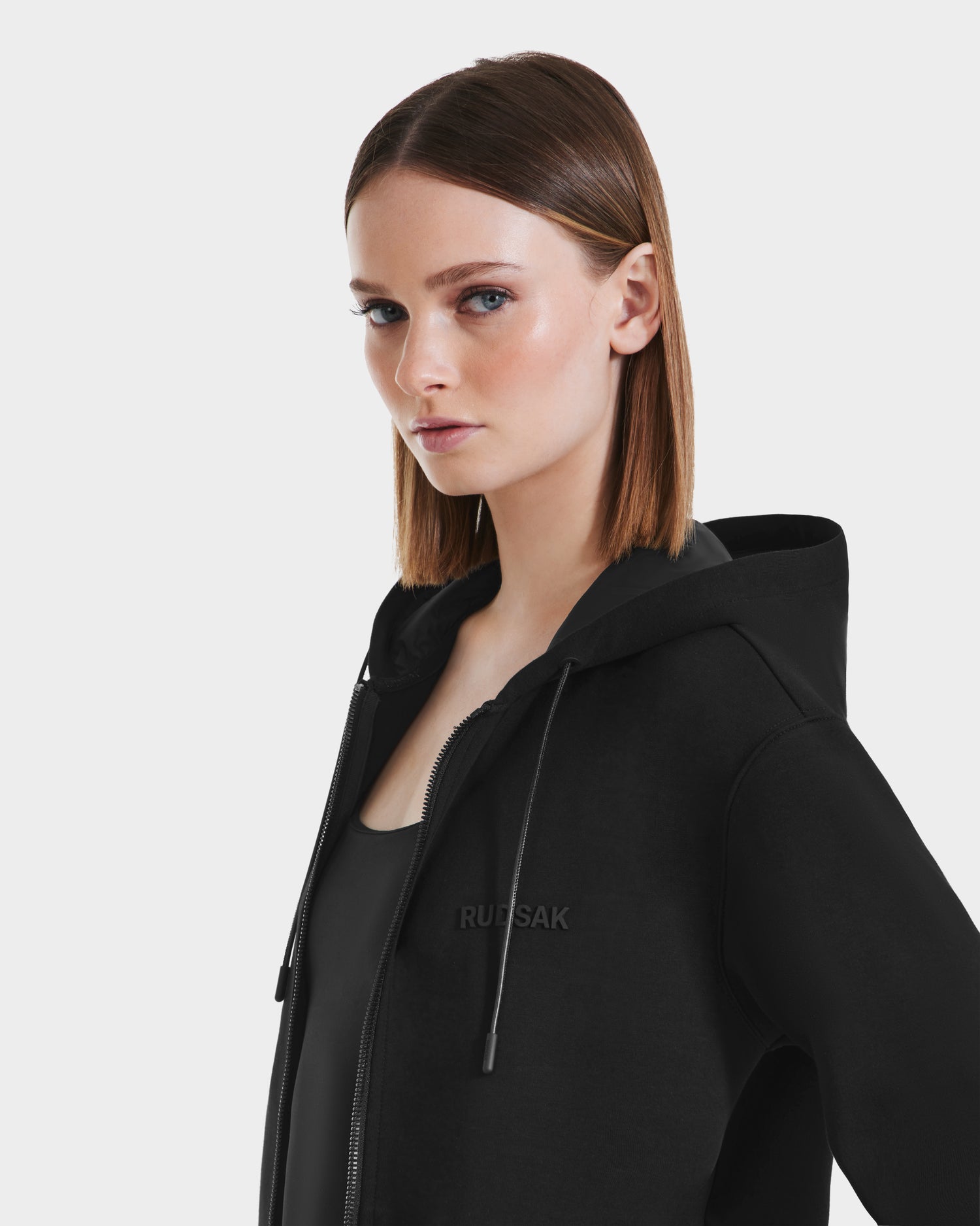 A woman with straight, shoulder-length brown hair wears the Rudsak LEVI - 9525552 Black W unisex zip-up hoodie with vegan leather trims over a black top, standing against a plain light background.
