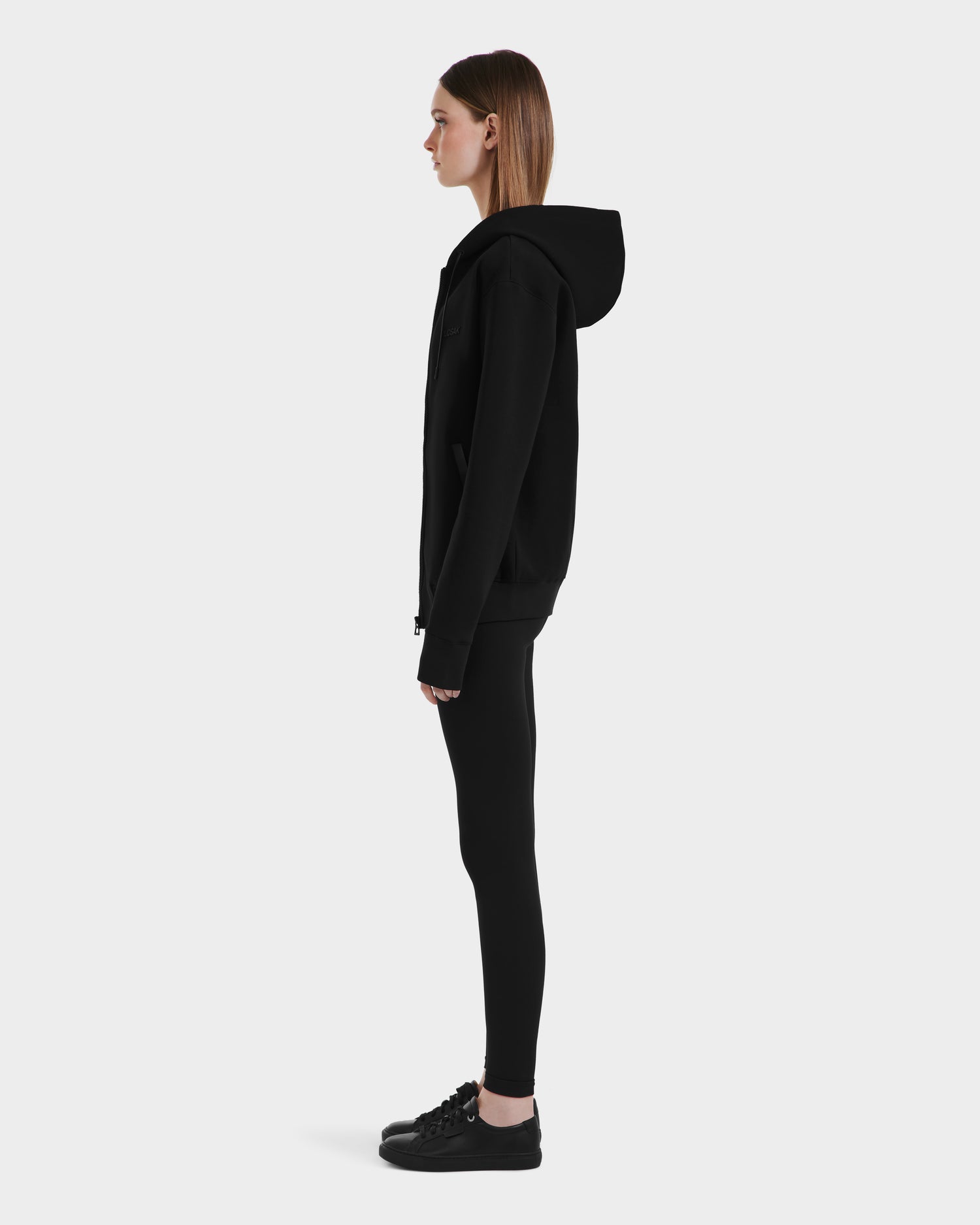 A woman in profile stands against a white background, wearing the Rudsak LEVI - 9525552 Black W hoodie, paired with black leggings and black sneakers.