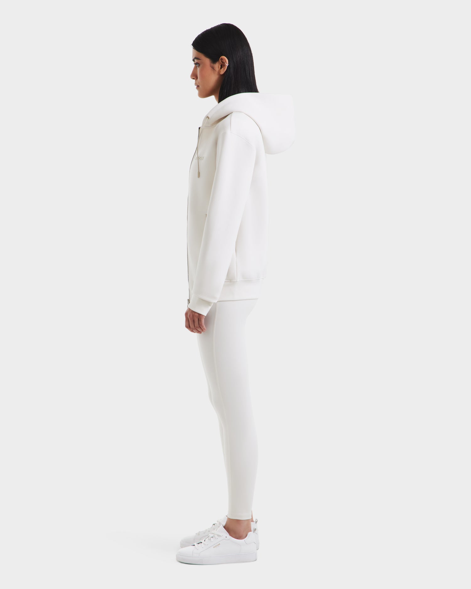 A woman stands in profile wearing the Rudsak LEVI - 9525552 Vanilla W unisex zip-up hoodie with vegan leather trims, paired with white leggings and sneakers, set against a plain white background.