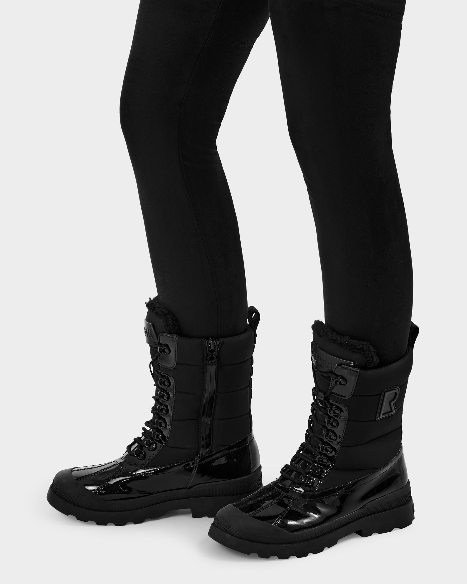 Someone in black leggings wears Rudsak LEX - 8224534 Black shiny, water-resistant winter boots with laces and thick soles, standing against a plain light background.