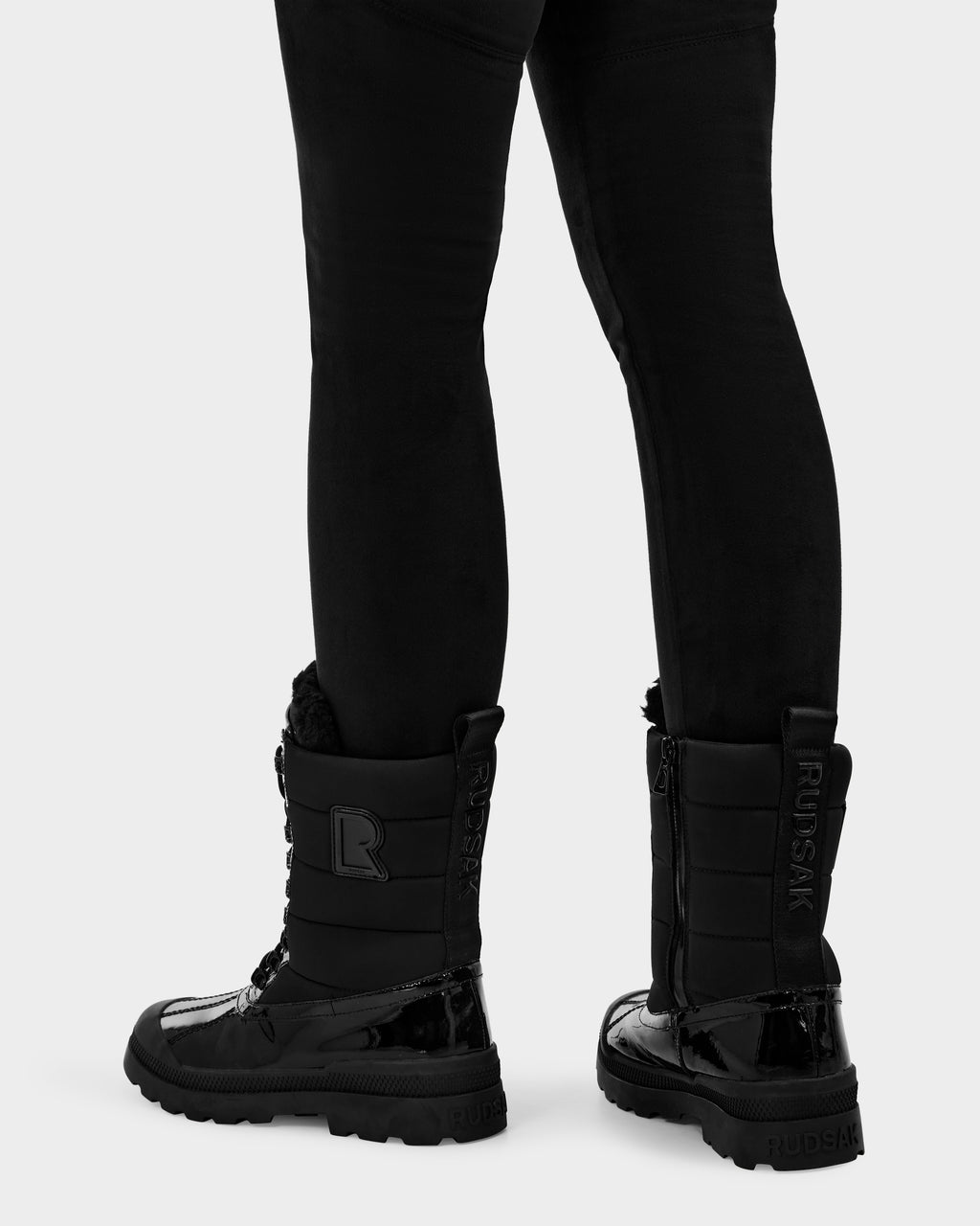 A person in black pants wears Rudsak’s LEX - 8224534 Black women’s winter boots with a shiny, water-resistant finish, wool lining, and visible branding on the side and back, standing on a light background.