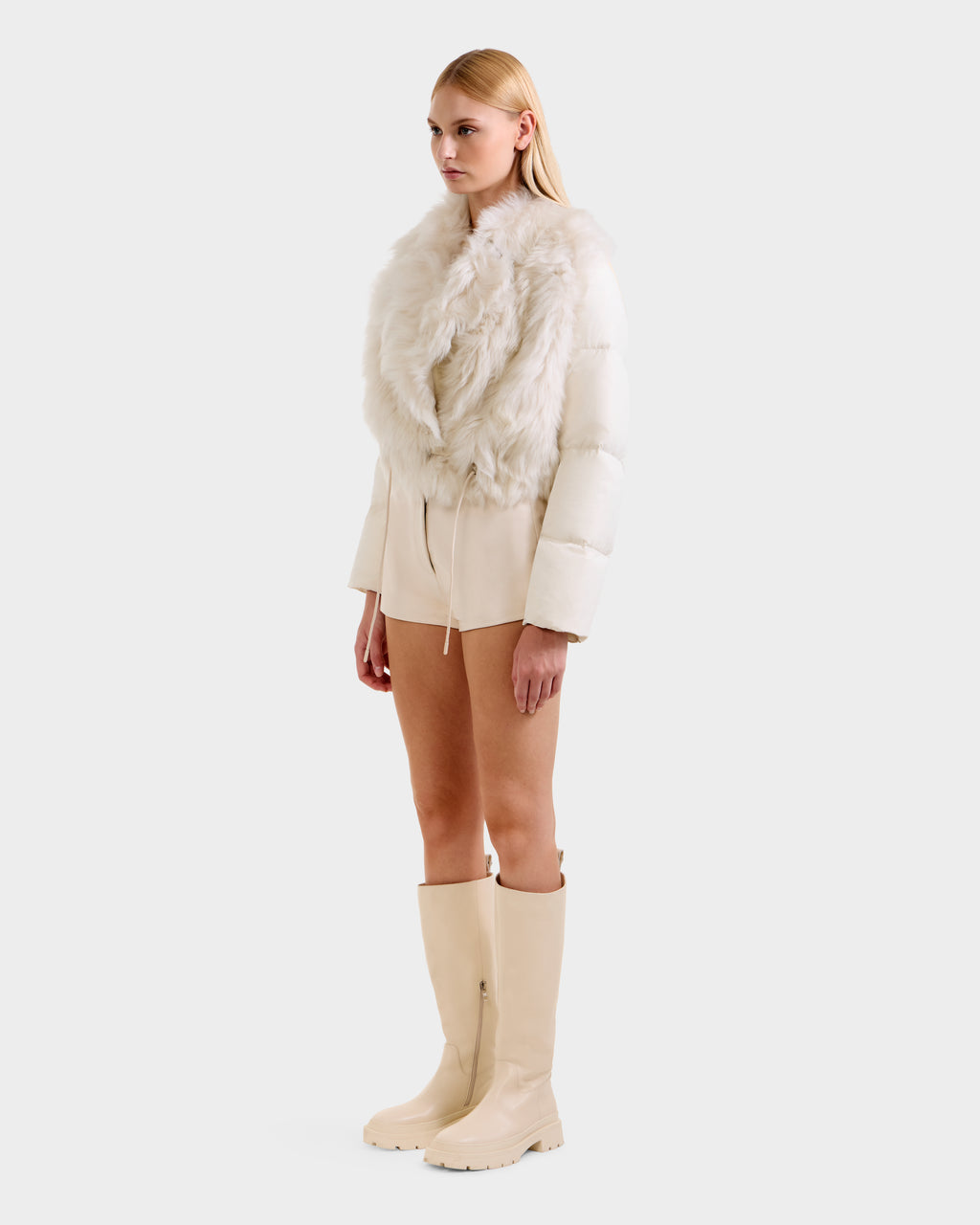 A woman with straight blonde hair and a neutral expression wears the Rudsak LILAH - 8125509 Vanilla cream shearling lamb jacket with puffer sleeves, paired with cream shorts and beige knee-high boots, posing against a plain white background.