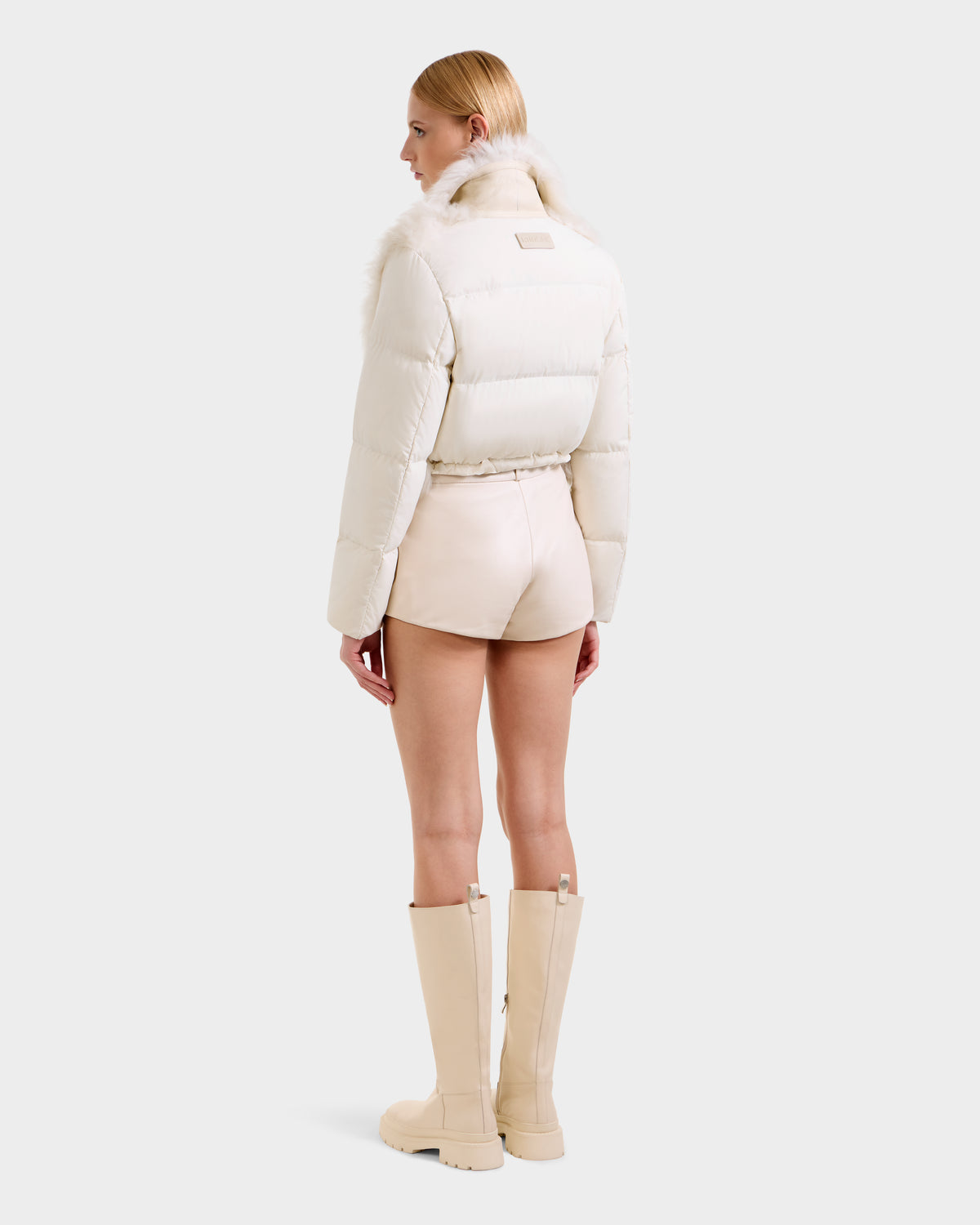 A person with long blonde hair faces away, wearing Rudsak's LILAH - 8125509 Vanilla cream-colored women’s cropped jacket with a fur collar, paired with matching high-waisted shorts and knee-high boots against a plain light background.