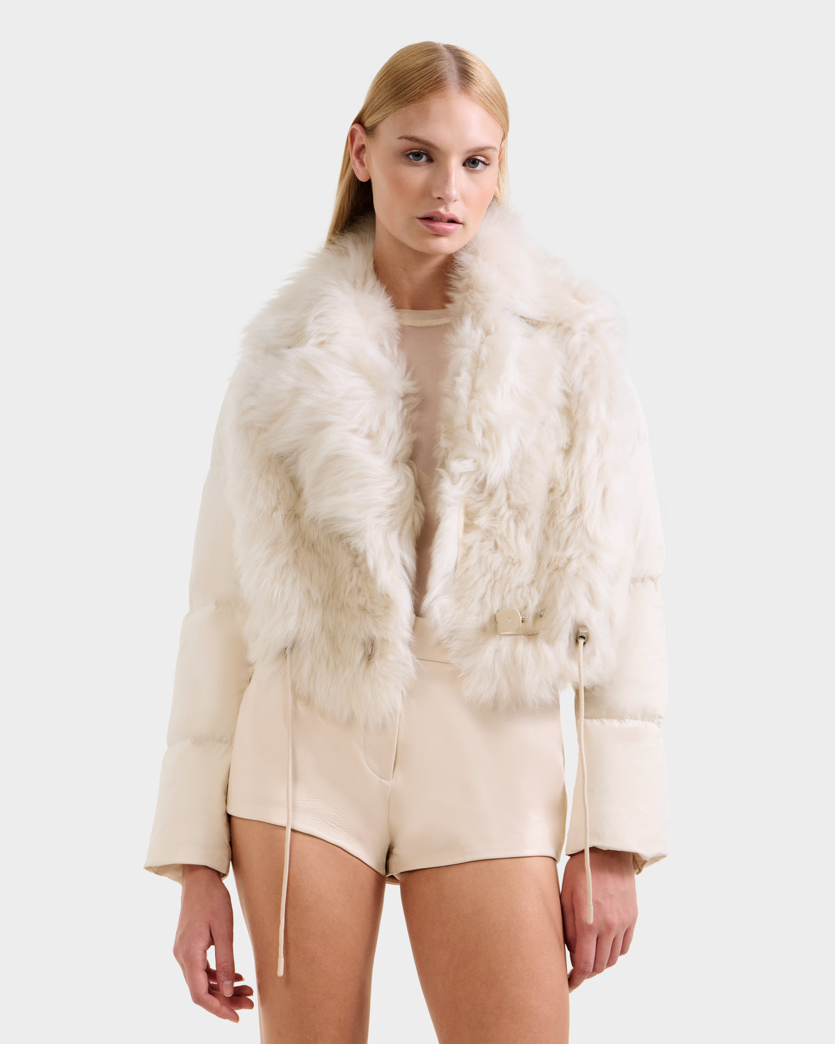 A woman with straight blonde hair models the Rudsak LILAH - 8125509 Vanilla women’s cropped jacket, paired with a beige top and matching high-waisted shorts, against a plain light background.