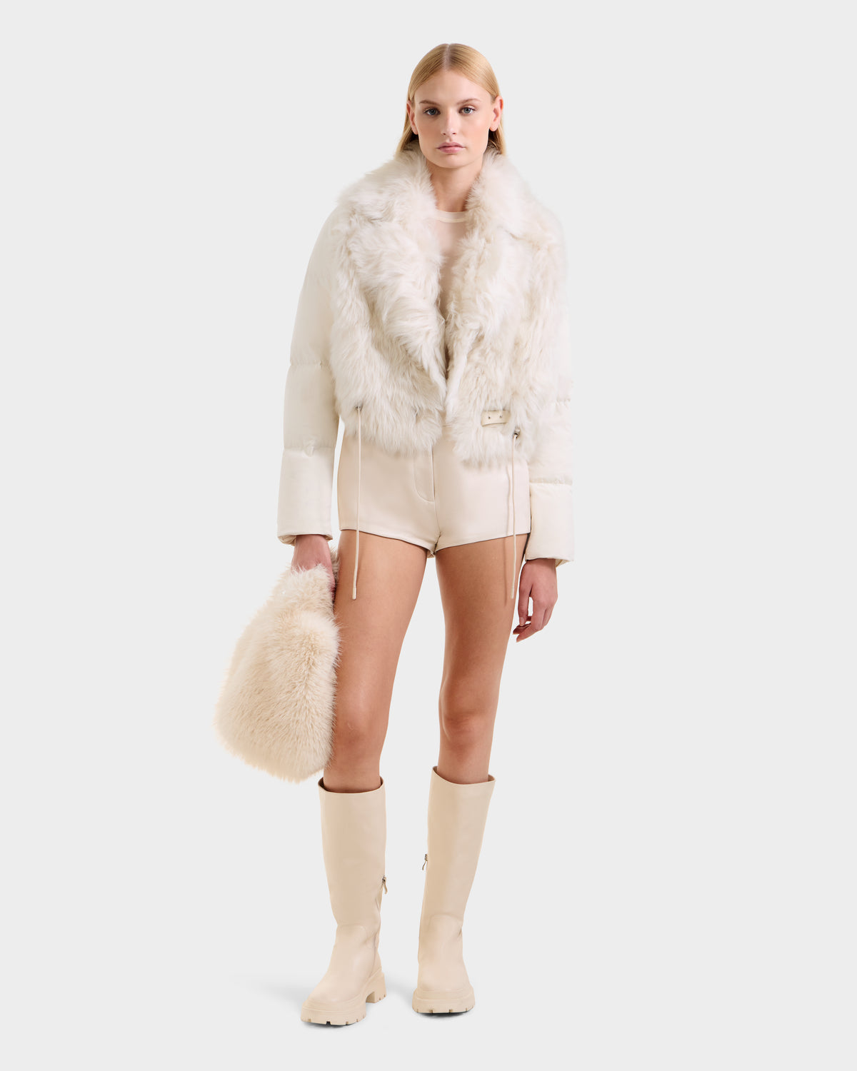 A woman wears the Rudsak LILAH - 8125509 Vanilla cream shearling lamb jacket with puffer sleeves, matching high-waisted shorts, cream knee-high boots, and holds a fluffy cream bag against a plain light gray background.