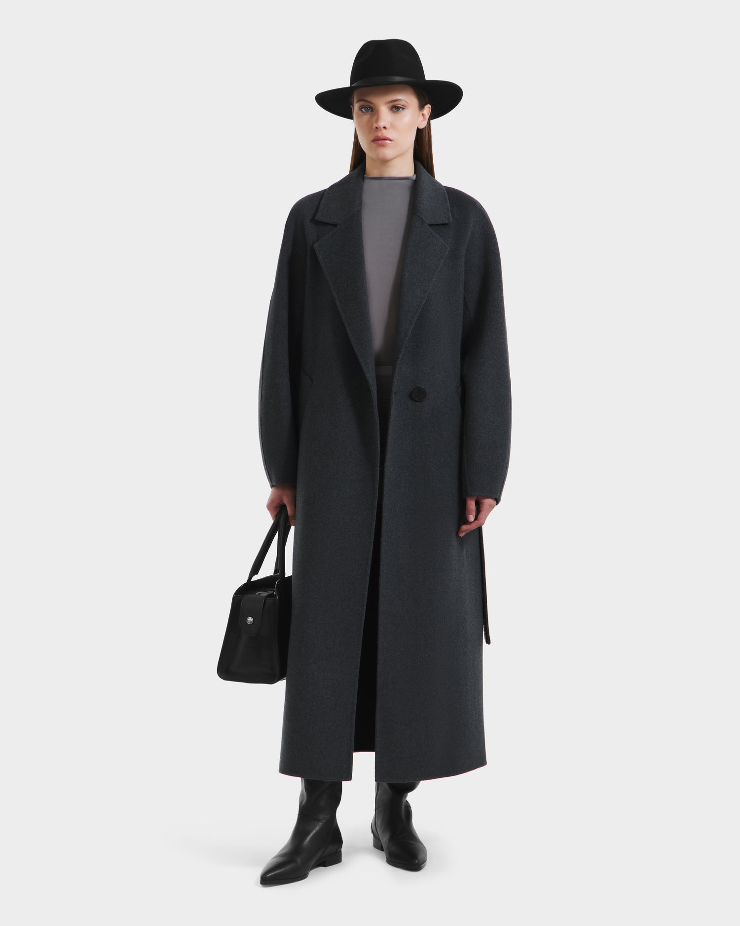A woman wearing the Rudsak LILIA - 8125634 Heather Charcoal wrap coat, along with a black wide-brim hat, gray top, black ankle boots, and holding a black handbag stands against a plain white background.