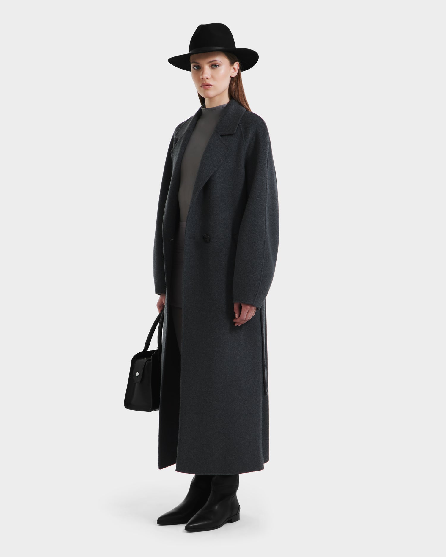 A woman wearing the Rudsak LILIA - 8125634 Heather Charcoal long wrap coat, paired with a black hat, gray top, black boots, and holding a black handbag stands against a plain white background.
