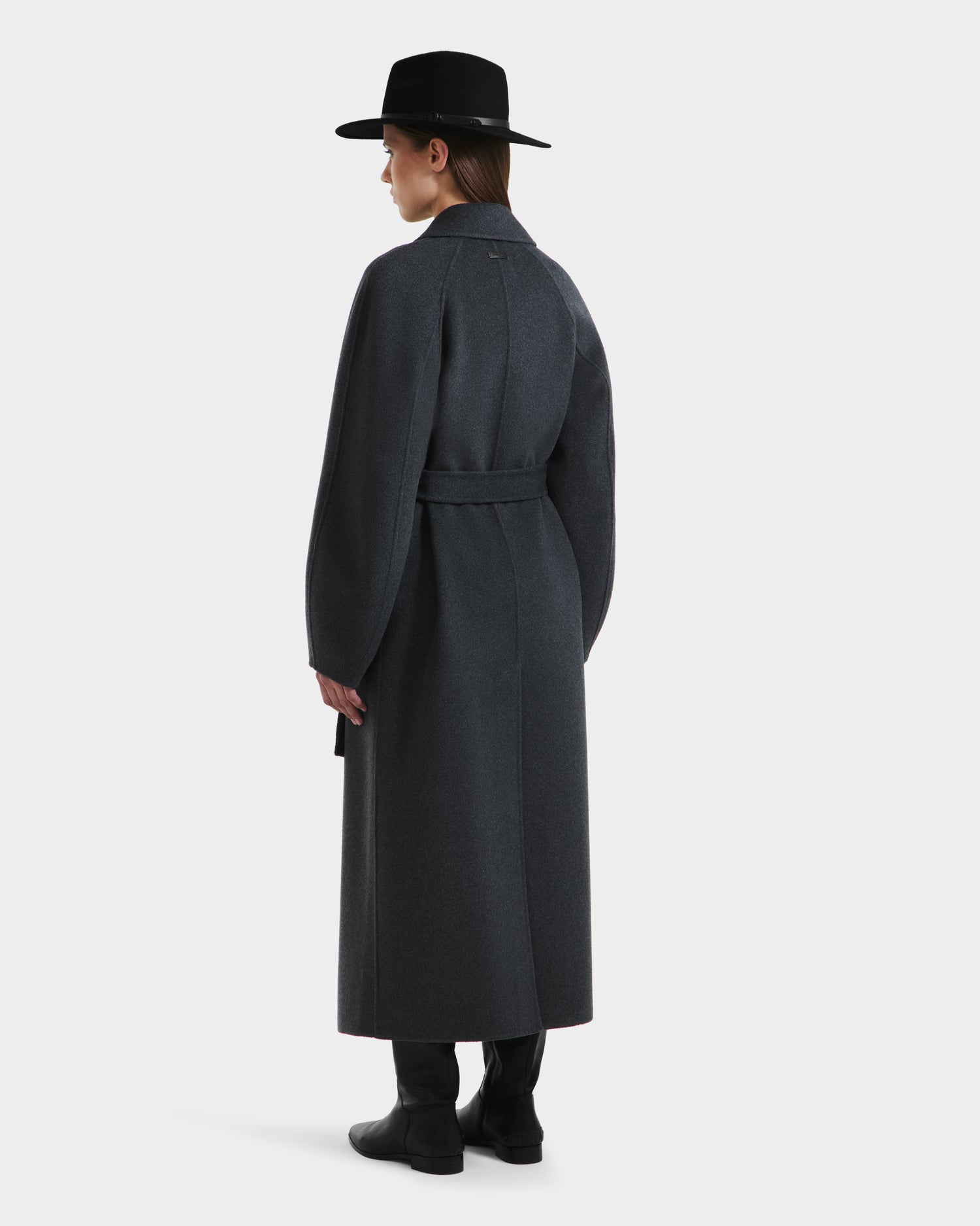 A person in the Rudsak LILIA - 8125634 Heather Charcoal womens long wrap coat with a belt, black boots, and a black wide-brimmed hat stands facing away from the camera against a plain light background.