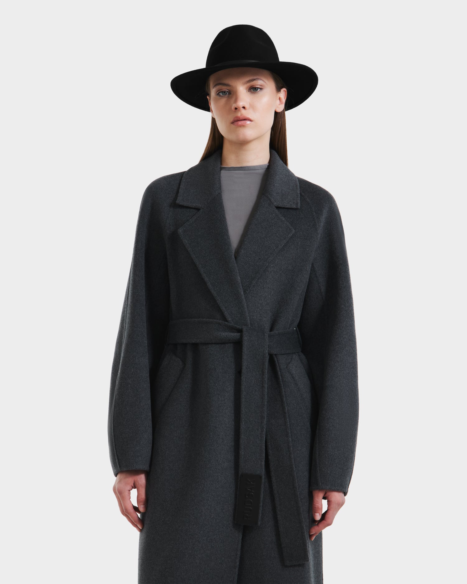 A woman stands against a plain light background wearing the Rudsak LILIA - 8125634 Heather Charcoal long wrap coat and a wide-brimmed black hat, looking straight ahead with a neutral expression.