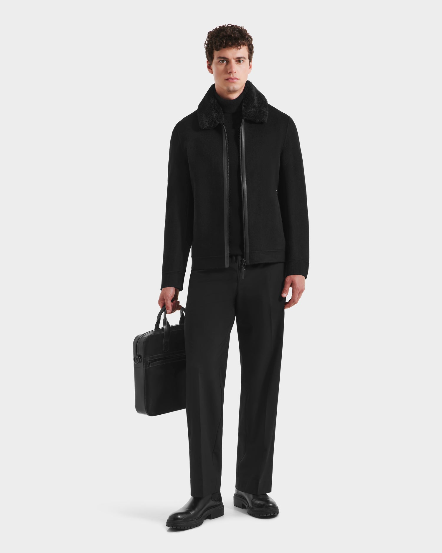 A man faces forward, wearing the Rudsak LINCOLN - 6125038 Black mens coat with removable faux fur collar, paired with a black turtleneck, pants, shoes, and holding a black briefcase against a plain light background.