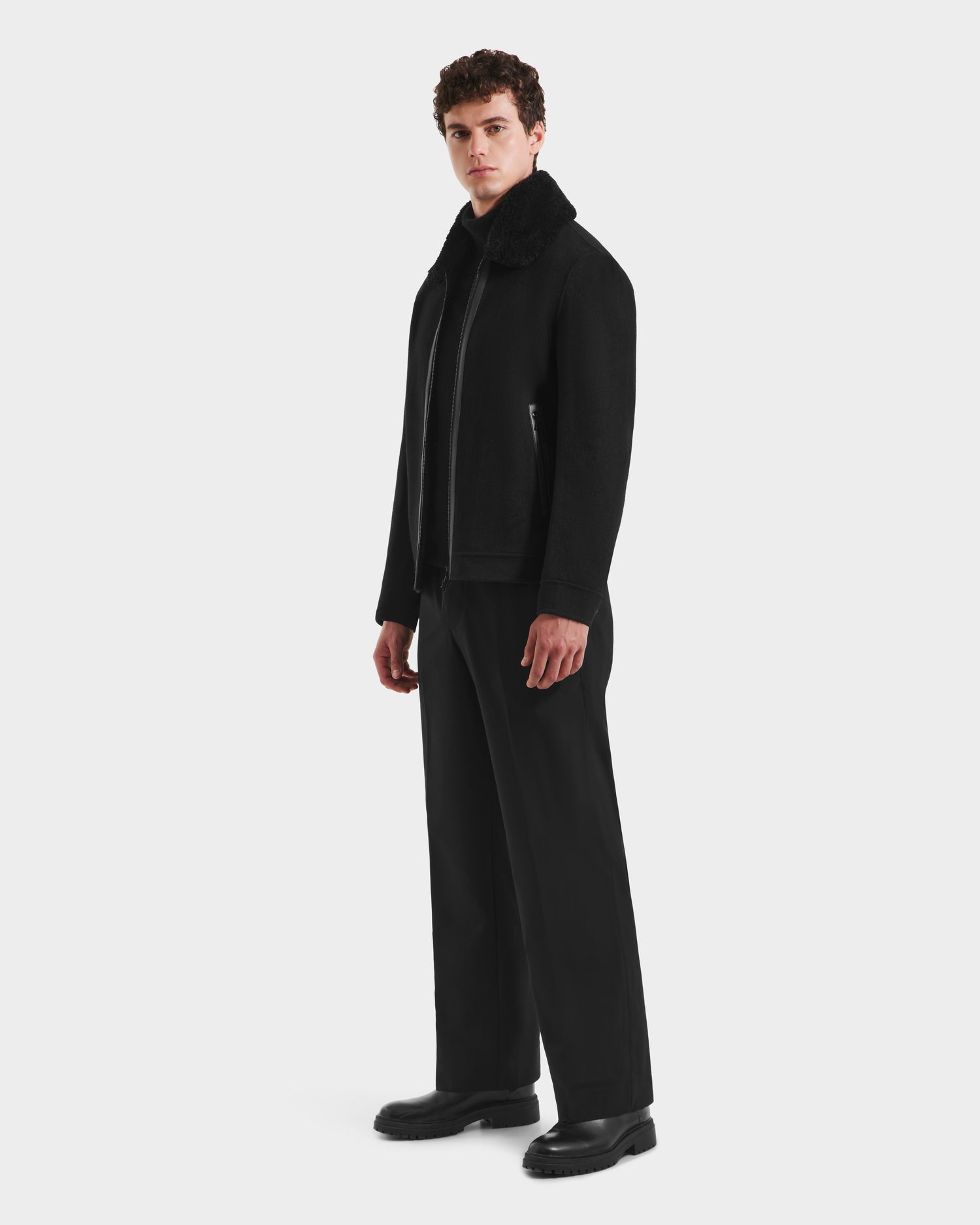 A man with curly hair stands against a white background, wearing the Rudsak LINCOLN - 6125038 Black mens coat with shearling collar, wide-leg black trousers, and black shoes, looking slightly to the side with a neutral expression.