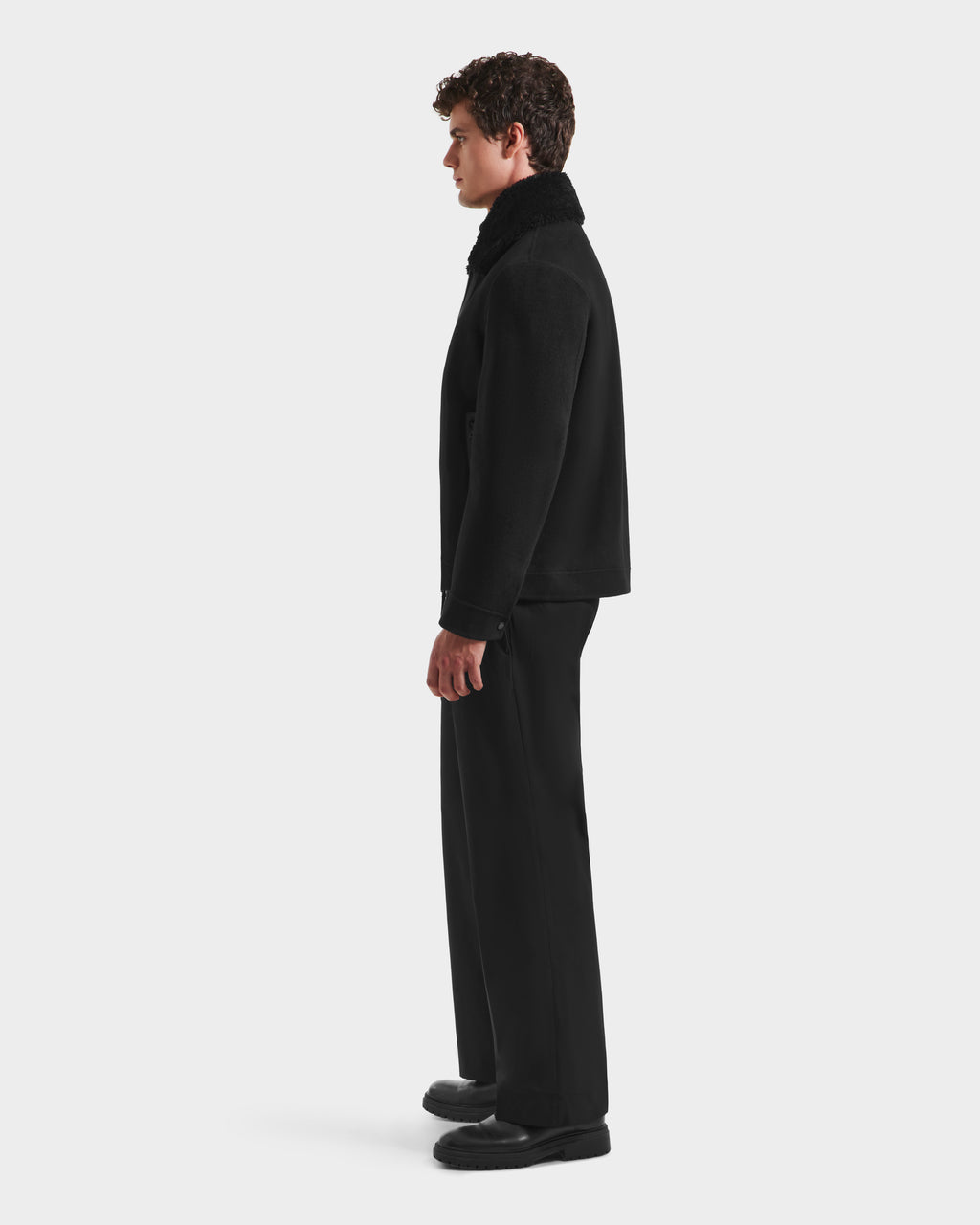 A man with curly brown hair stands in profile against a white background, wearing Rudsaks LINCOLN - 6125038 Black coat featuring a removable faux fur collar, paired with black trousers and shoes.