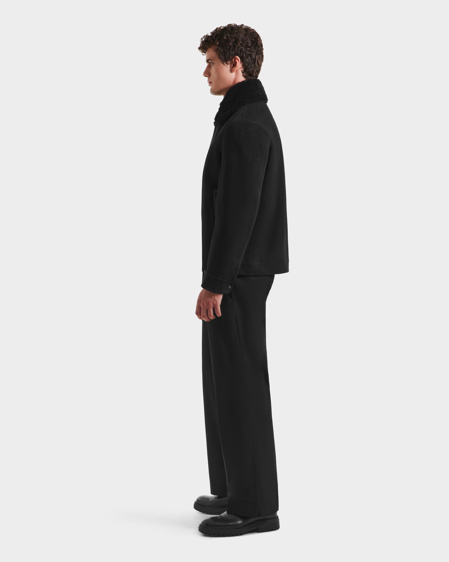 A man with curly brown hair stands in profile against a white background, wearing Rudsaks LINCOLN - 6125038 Black coat featuring a removable faux fur collar, paired with black trousers and shoes.