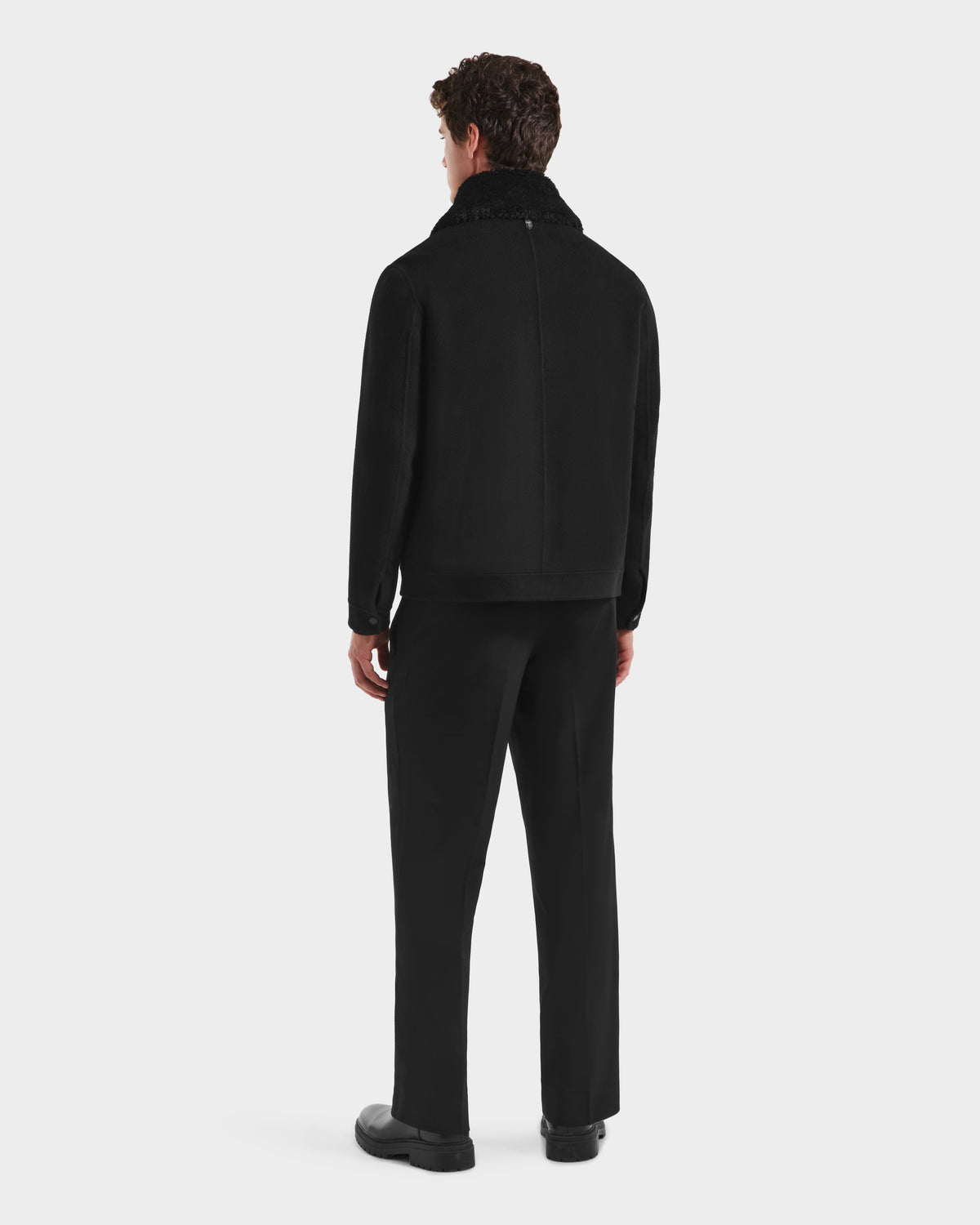 A man with curly hair stands facing away, wearing the Rudsak LINCOLN - 6125038 Black mens coat with a removable faux fur collar, paired with black pants and shoes, against a plain white background.