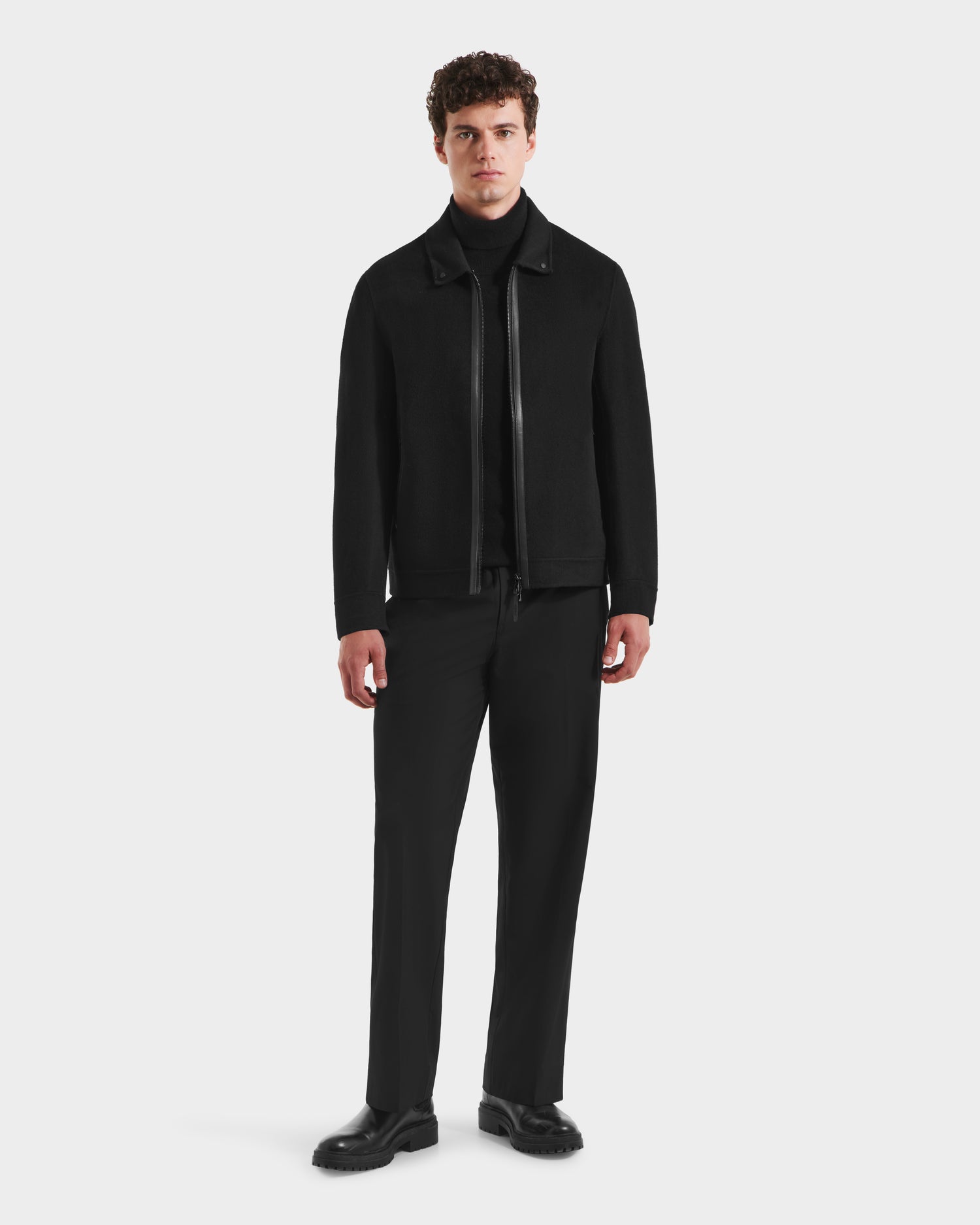 A man with short curly hair faces forward, wearing the Rudsak LINCOLN - 6125038 Black mens coat with a removable faux fur collar, paired with black pants and shoes, set against a plain white background.